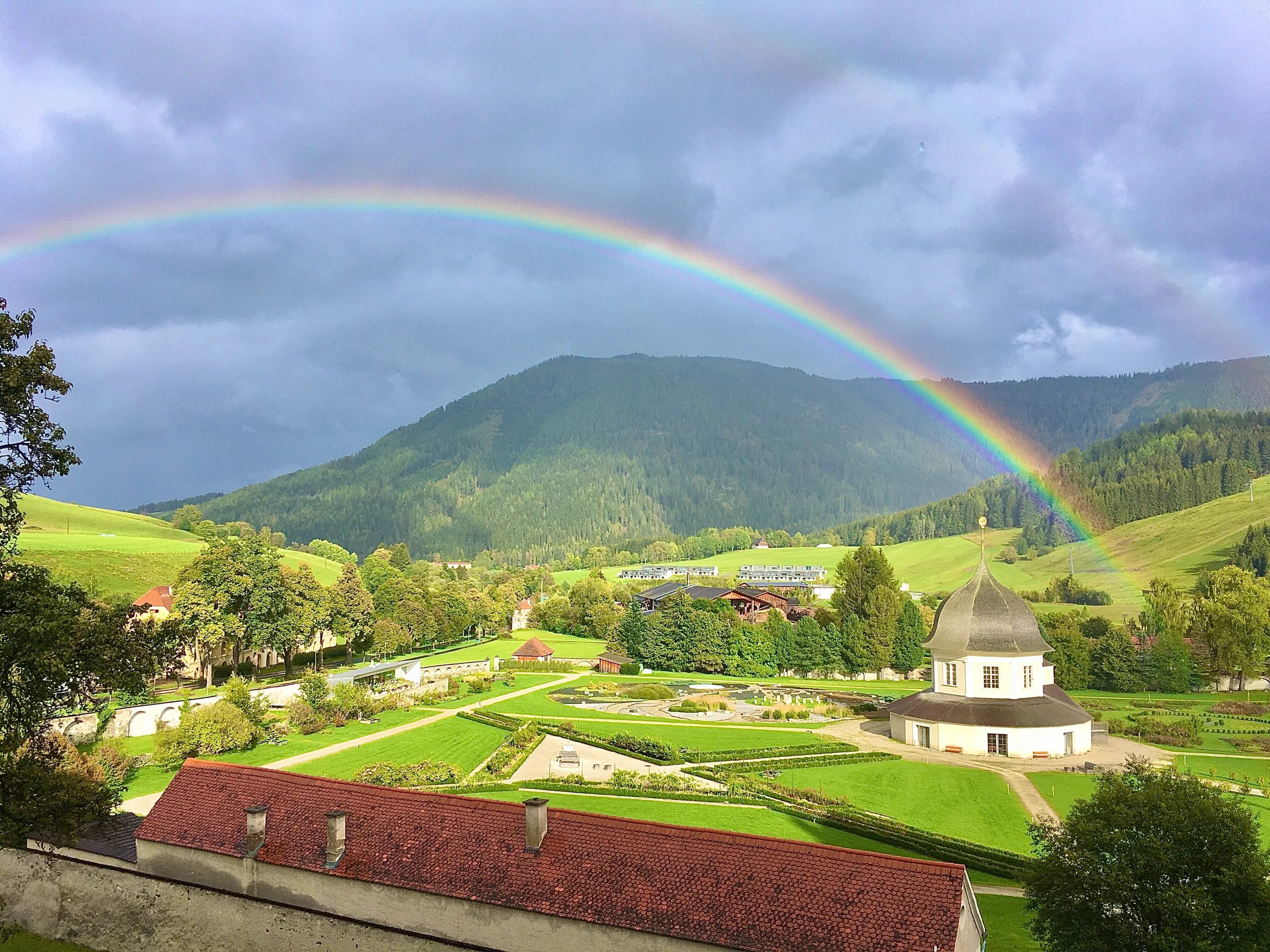 Regenbogen über den bewaldeten Bergen, darunter der Stiftsgarten mit Pavillon