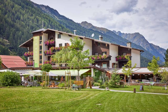 Außenansicht des Hotels mit blühenden Balkonen, Terrasse und Garten vor Bergkulisse
