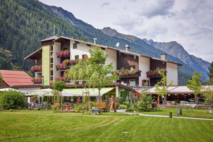 Außenansicht des Hotels mit blühenden Balkonen, Terrasse und Garten vor Bergkulisse