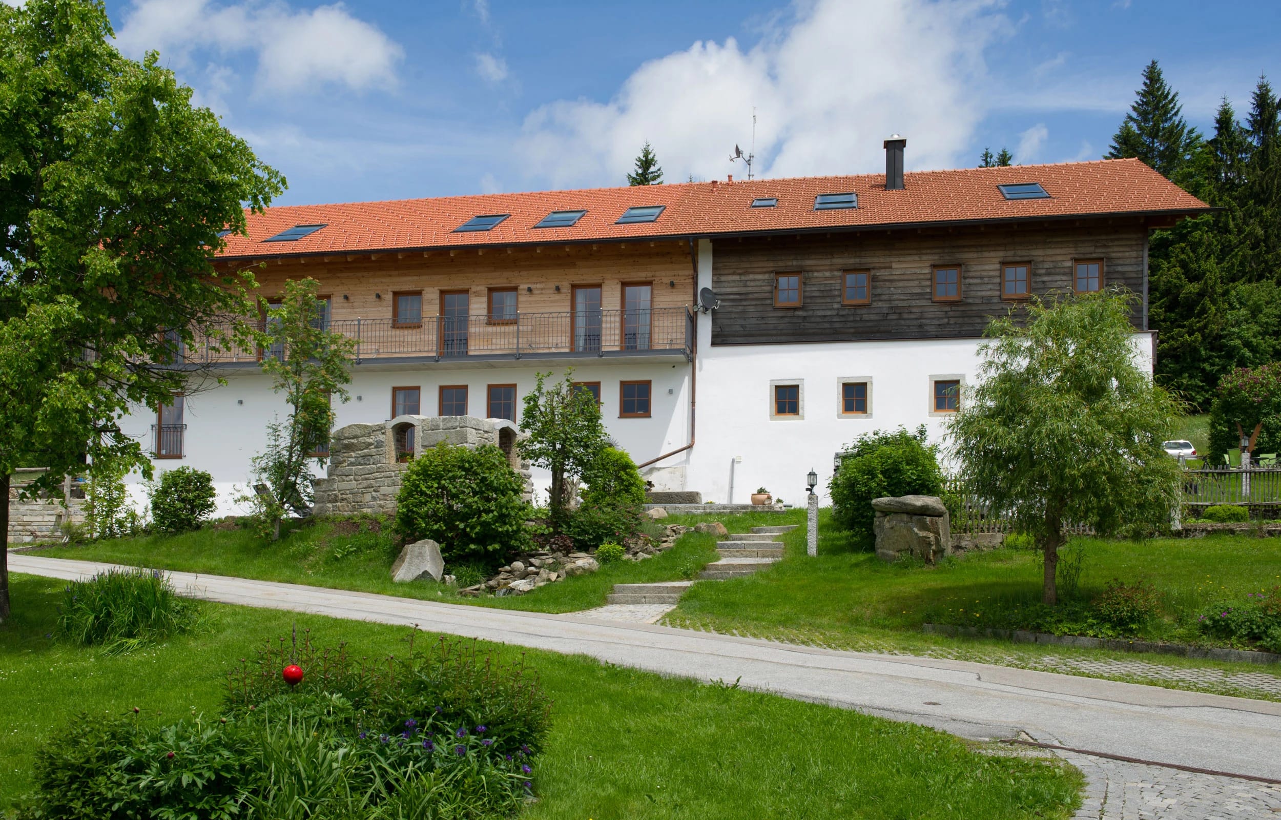 Langgestrecktes Gebäude mit weißer Fassade, holzverkleidetem Obergeschoss und rotem Ziegeldach, davor Wiese und Zufahrt