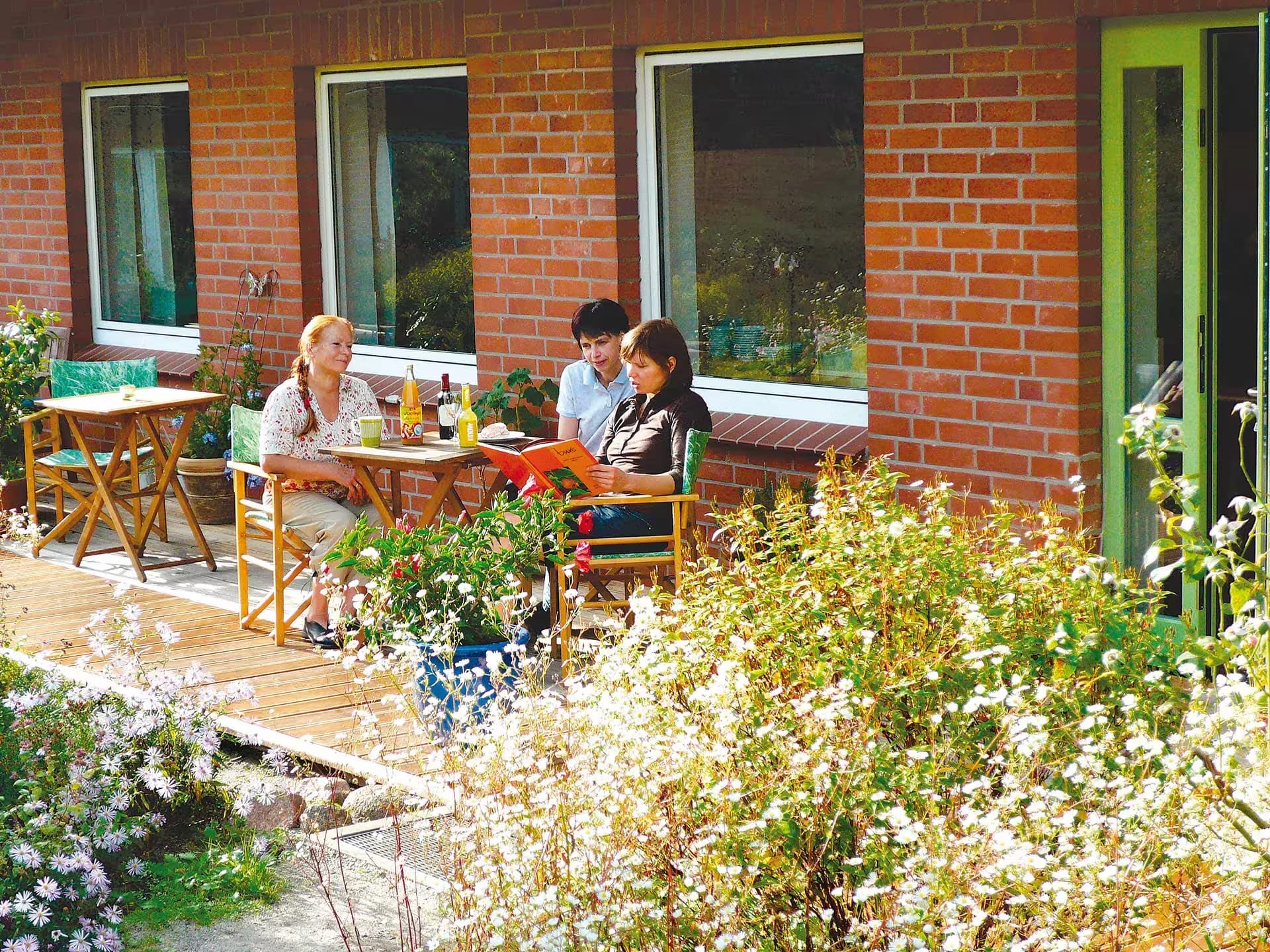 Sonnige Holzterrasse mit Sitzplätzen vor der Backsteinfassade, umgeben von Blumenbeeten