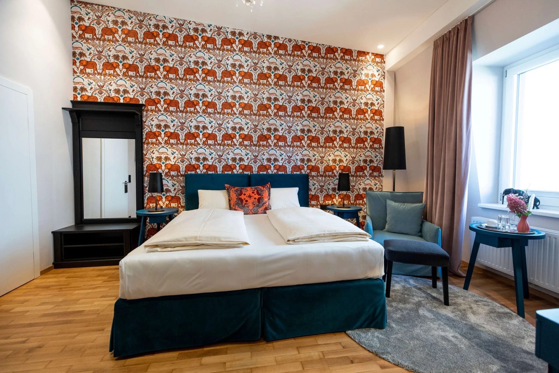 Zimmer mit orange gemusterter Tapete, türkisfarbenem Bett, Sessel und Fenster mit Beistelltisch