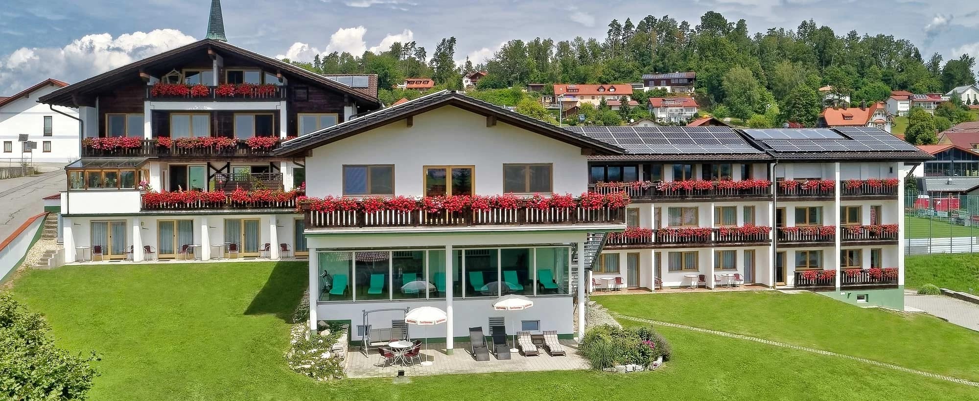 Hotelgebäude mit weißer Fassade, holzverkleideten Balkonen mit rotem Blumenschmuck und verglaster Terrasse mit Liegestühlen auf der Wiese