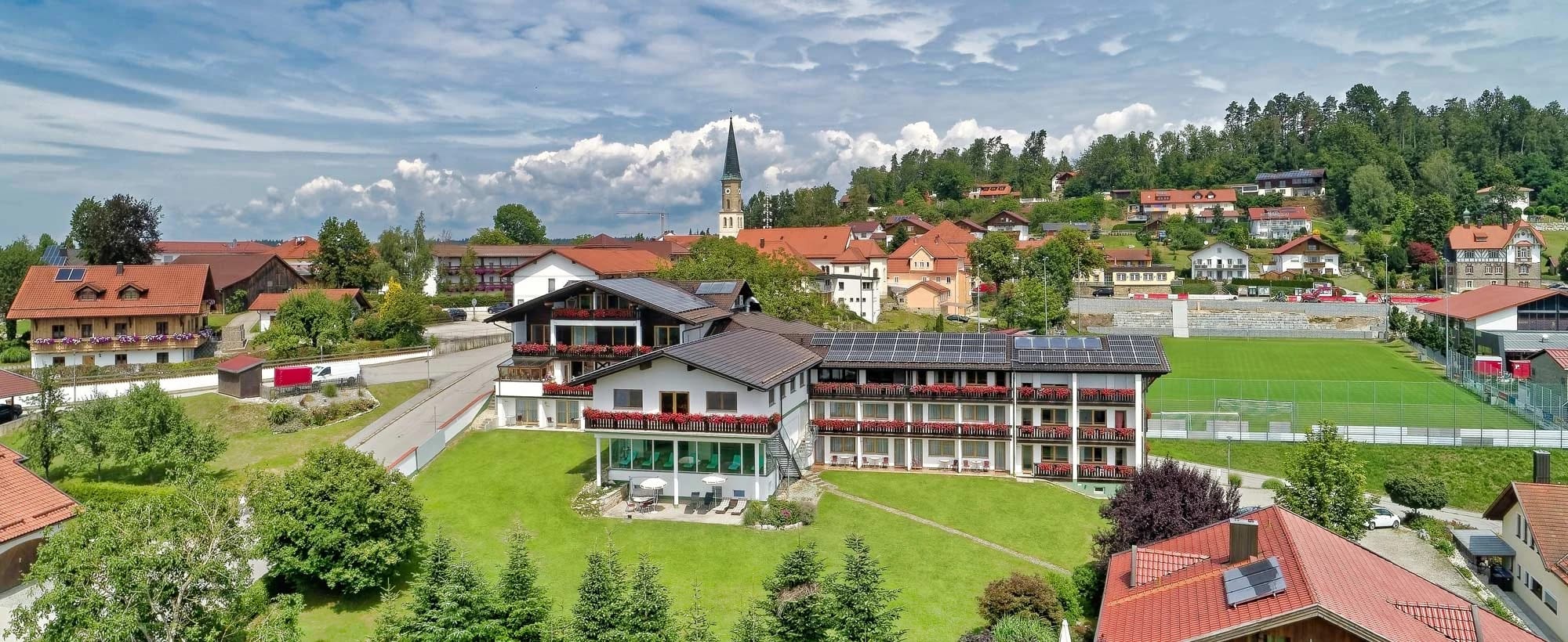 Luftaufnahme des Hotels inmitten von Büchlberg mit Wiesen, Kirchturm und bewaldeten Hügeln