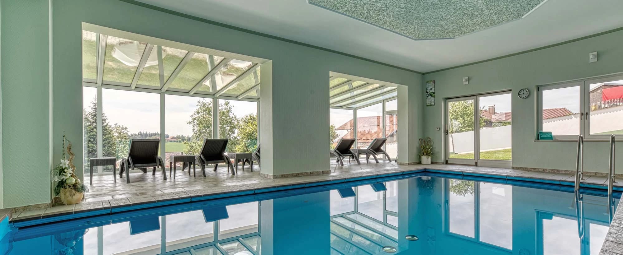 Hallenbad mit blauem Becken, großer Fensterfront und Liegestühlen am Beckenrand