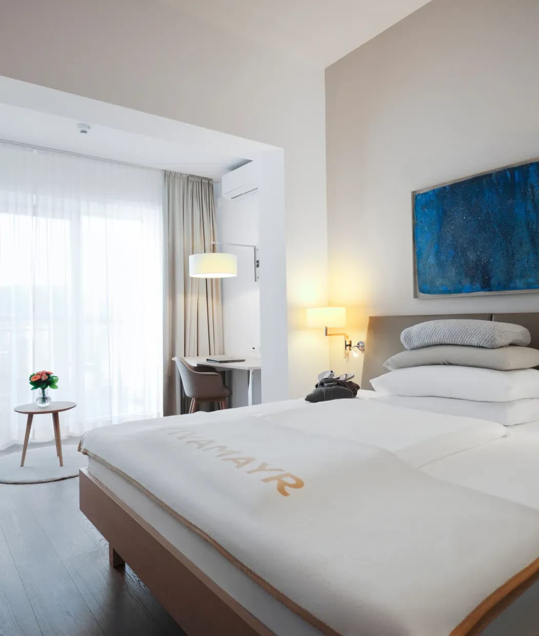 Zimmer mit breitem Doppelbett, Leselampen, blauem Bild über dem Kopfteil und Fenstertür zum Balkon