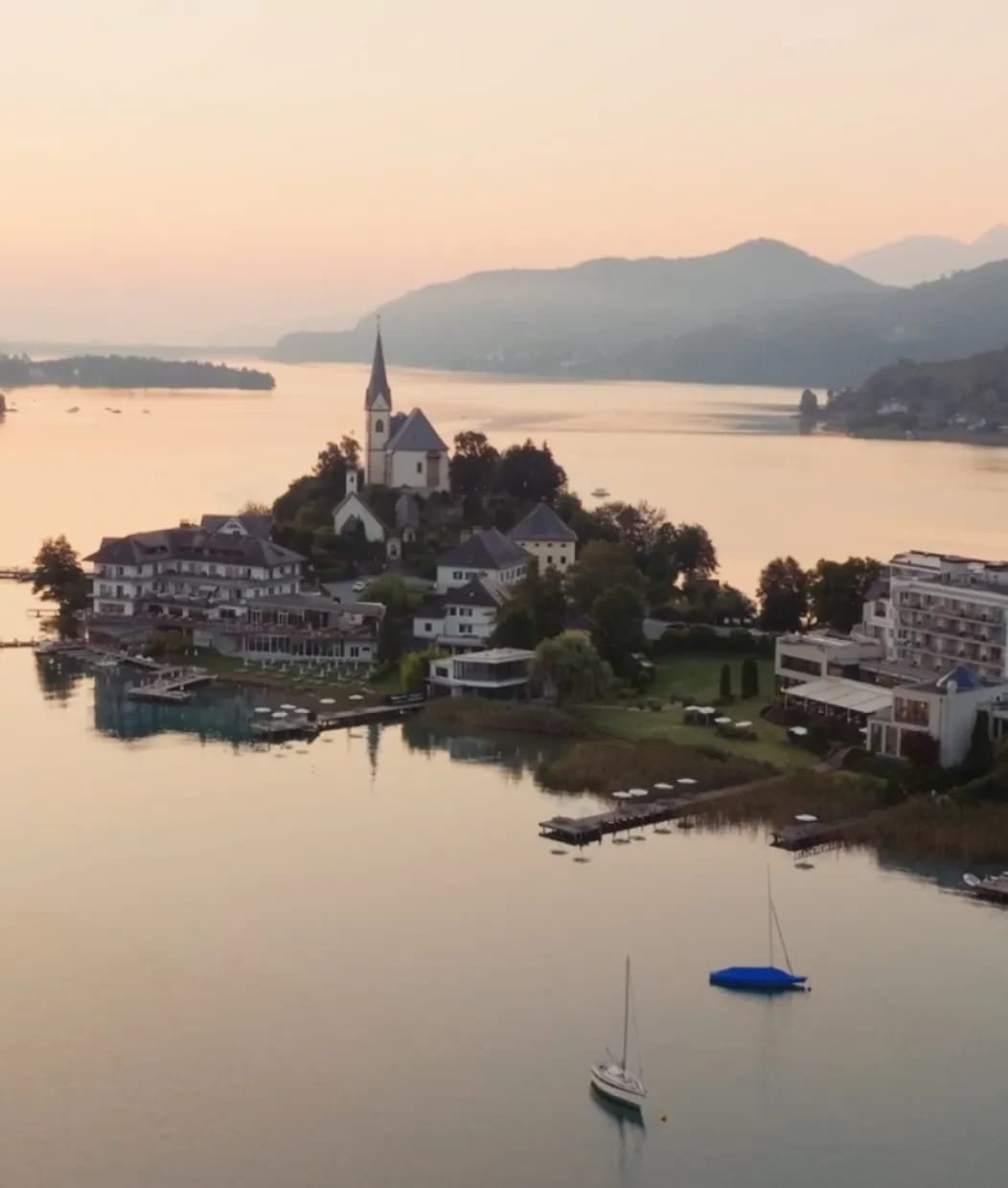 Luftaufnahme der Halbinsel von Maria Wörth mit Kirchturm, Segelbooten und dem Wörthersee im Morgenlicht