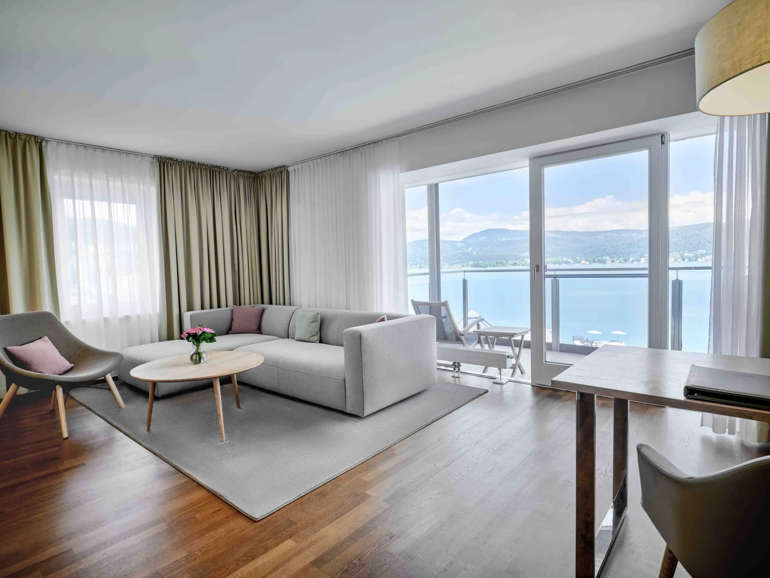 Wohnbereich einer Suite mit grauem Sofa, Sesseln und bodentiefen Fenstern zum Balkon mit Seeblick