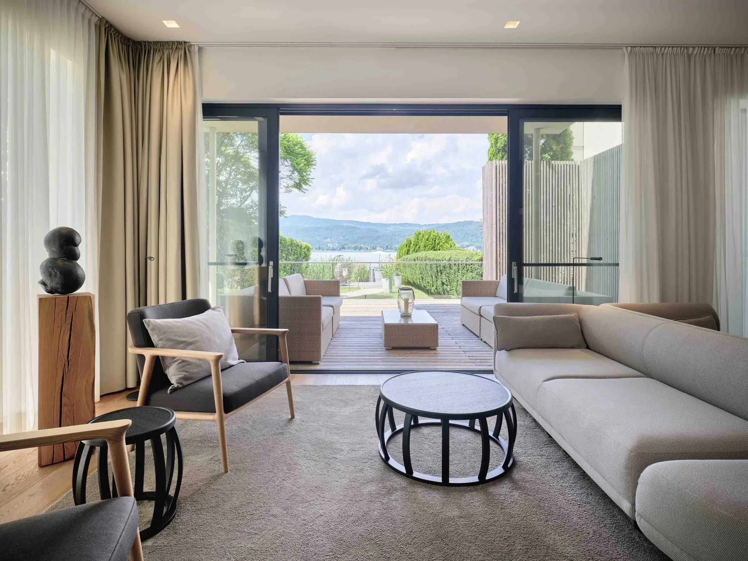 Wohnzimmer mit langer Sofalandschaft, Sesseln und runden Beistelltischen, offene Glasfront zur Terrasse mit Seeblick