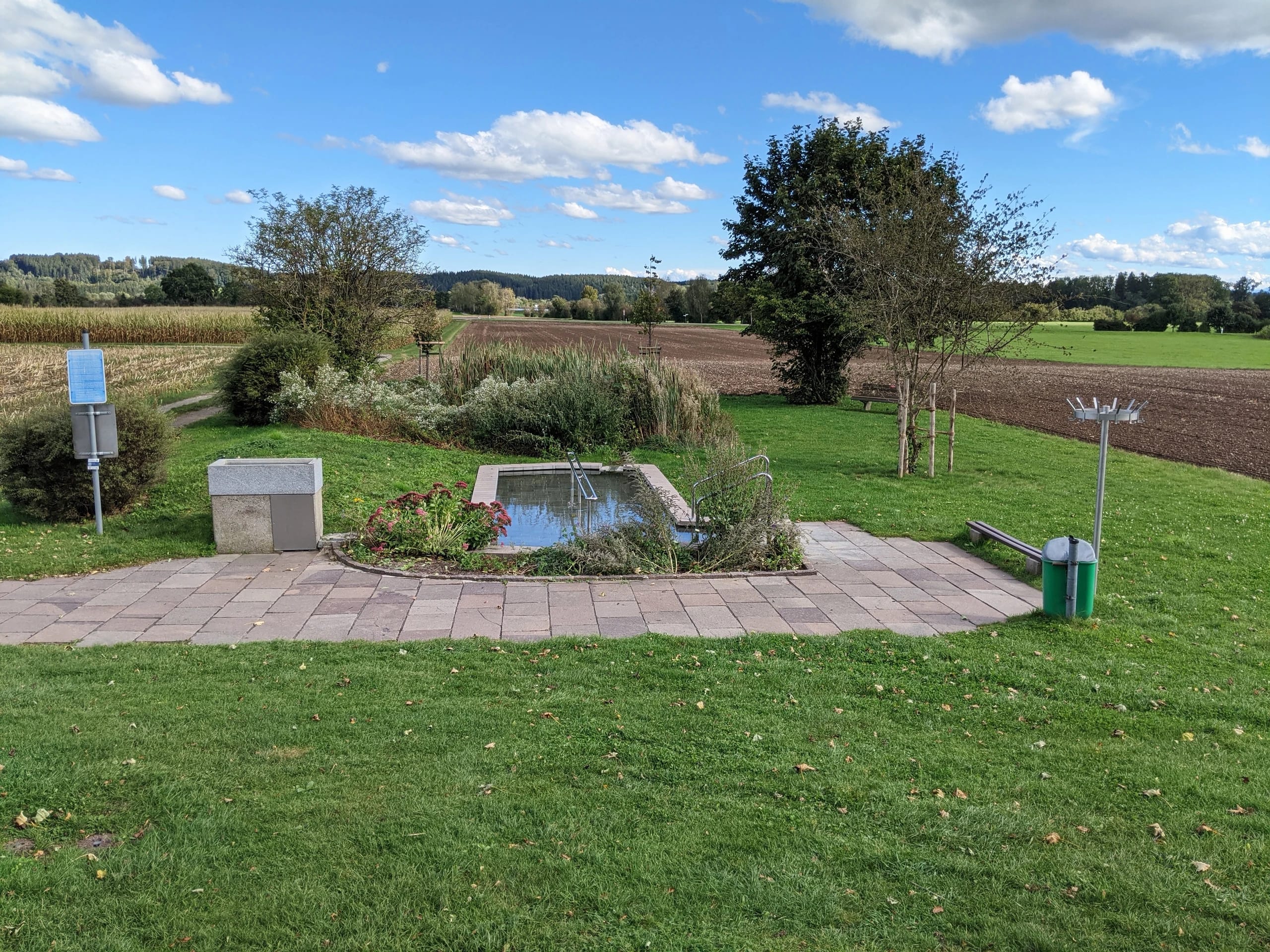Kneipp-Wassertretbecken mit Haltestange im Freien, umgeben von Wiese, Feldern und Bäumen