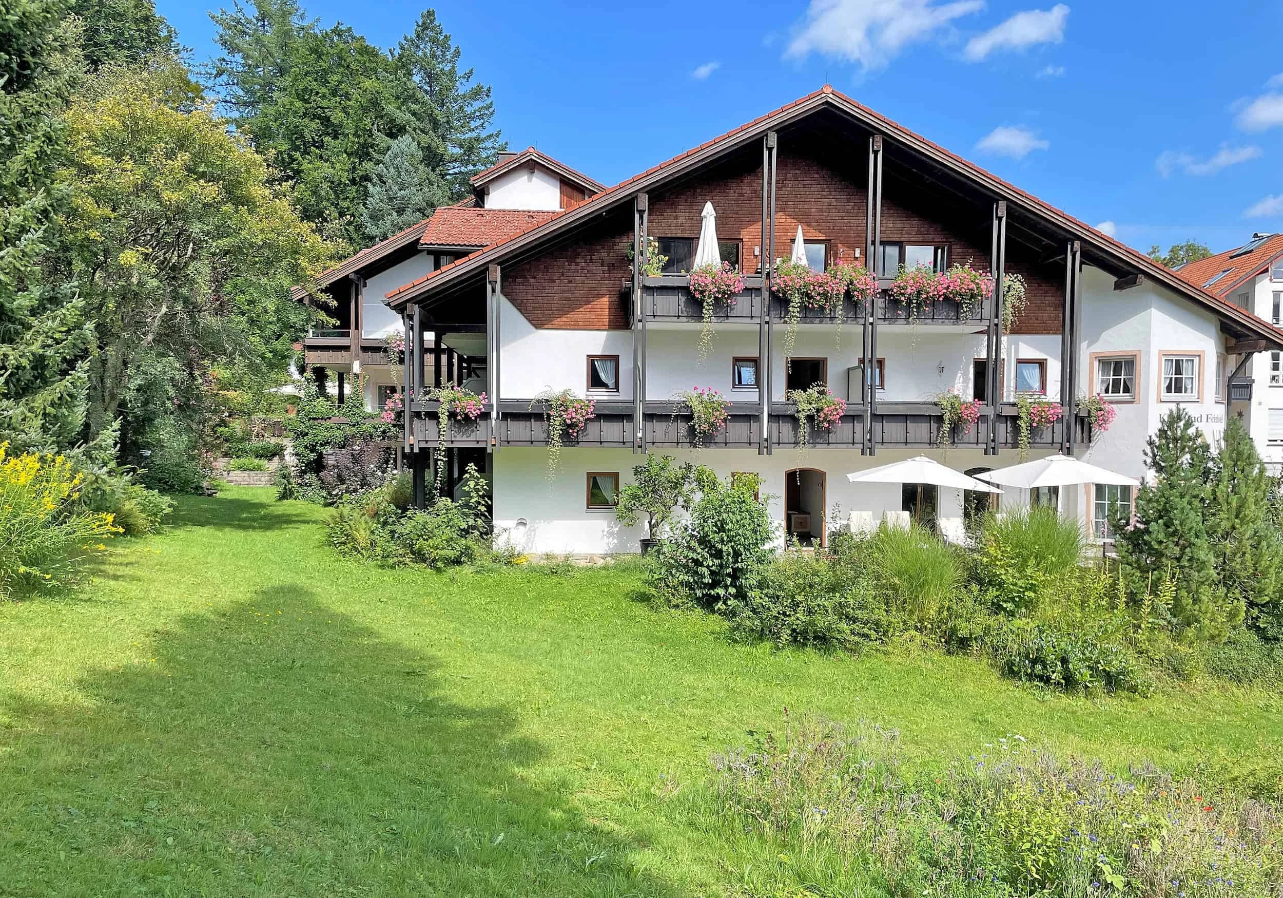 Außenansicht des Hauses mit Balkonen, Blumenkästen und Sonnenschirmen, davor eine Wiese bei blauem Himmel