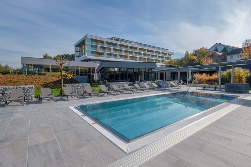 Außenansicht des Hotels mit Außenpool, Sonnenliegen und herbstlichen Bäumen bei blauem Himmel