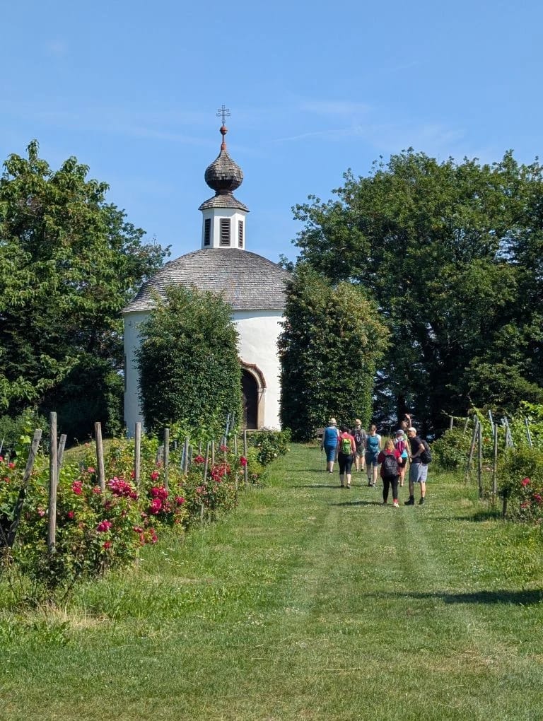 Kleine Kapelle am Ende eines von Rosen gesäumten Wegs, im Hintergrund eine Gruppe