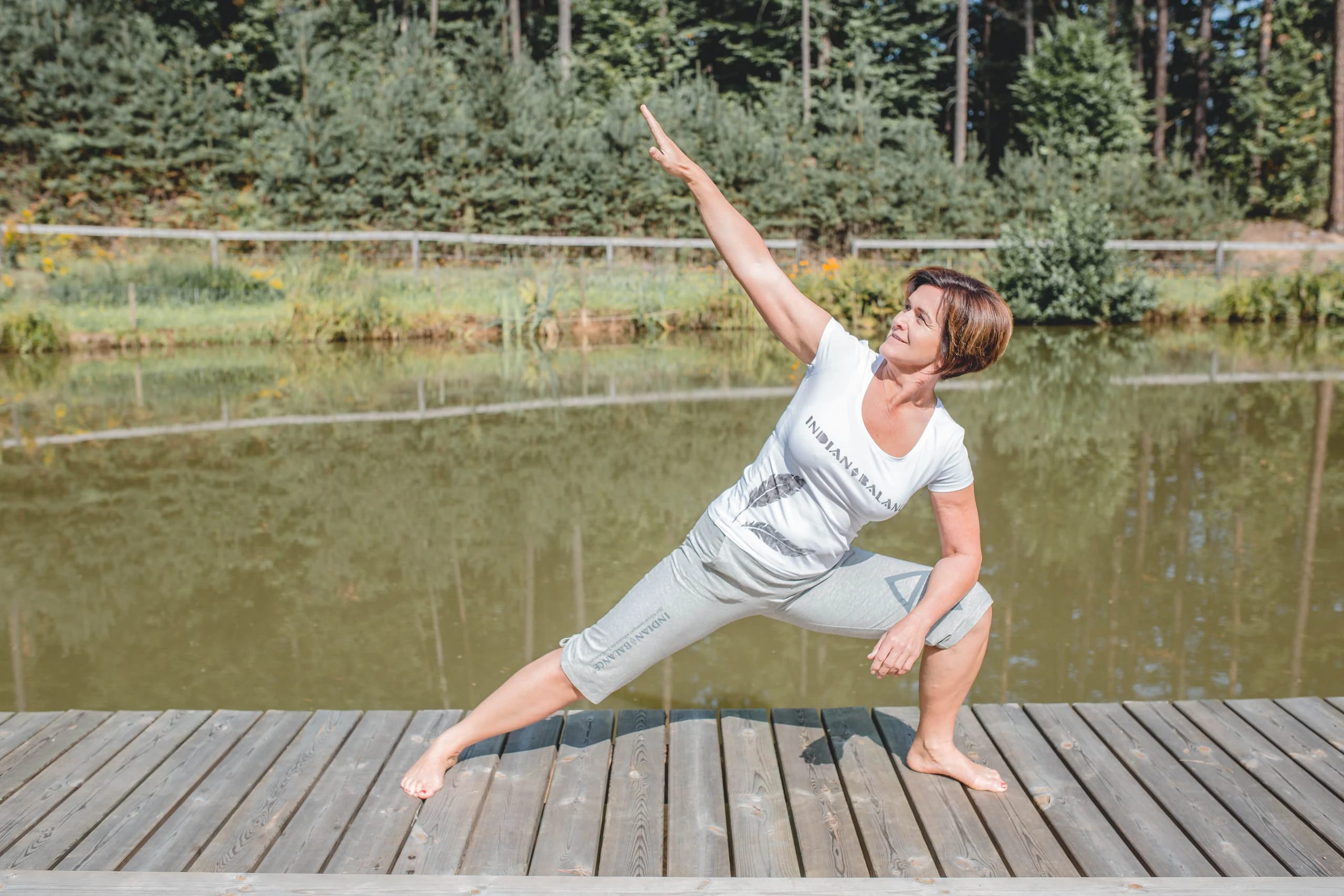 Frau in einer Yoga-Haltung mit ausgestrecktem Arm auf einem Holzsteg am Wasser