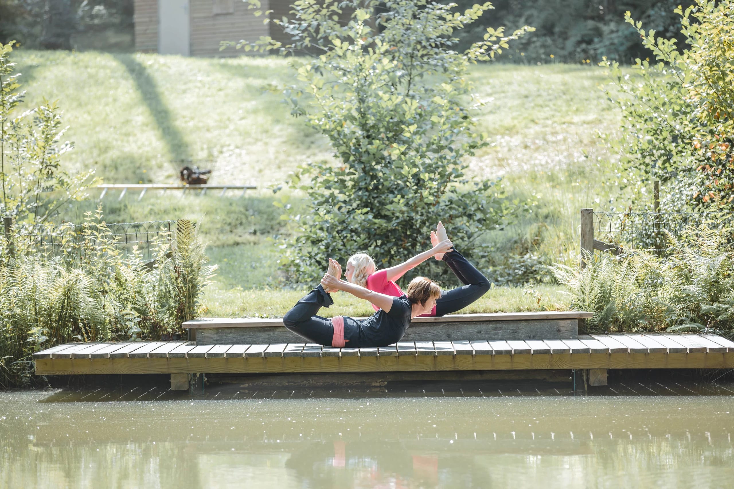 Zwei Frauen bei einer Partner-Yoga-Übung auf einem Holzsteg am Wasser