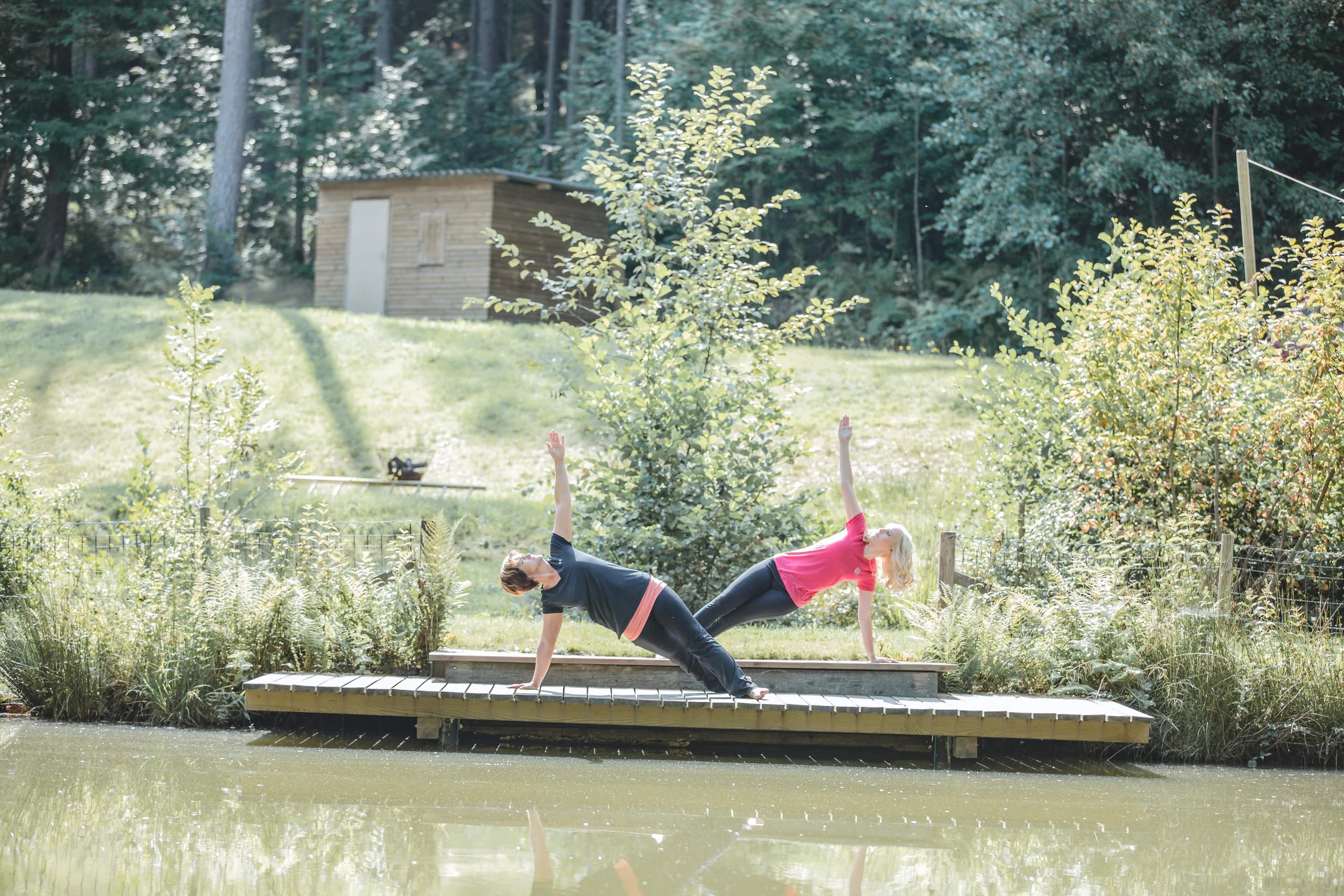 Zwei Frauen in einer Seitstütz-Yoga-Haltung auf einem Holzsteg am Wasser