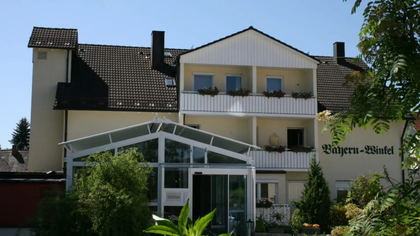 Außenansicht des Hauses mit gläsernem Wintergarten-Eingang, Balkonen mit Blumenkästen und Grün davor