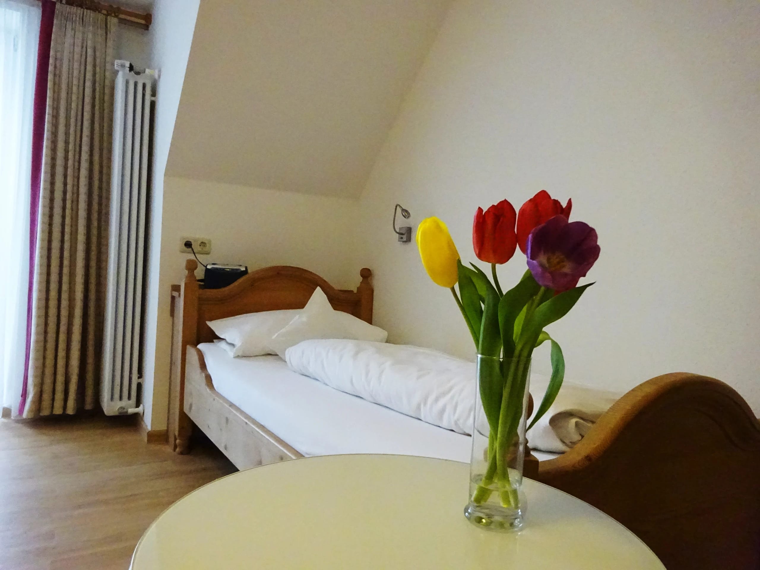 Einzelzimmer mit Holzbett, Vorhang am Fenster und rundem Tisch mit Tulpenstrauß im Vordergrund