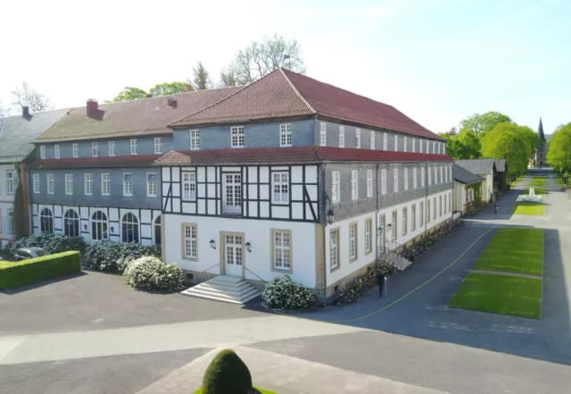 Herrenhaus mit rotem Ziegeldach und Fachwerkgiebel, davor gepflasterter Vorplatz mit blühenden Sträuchern