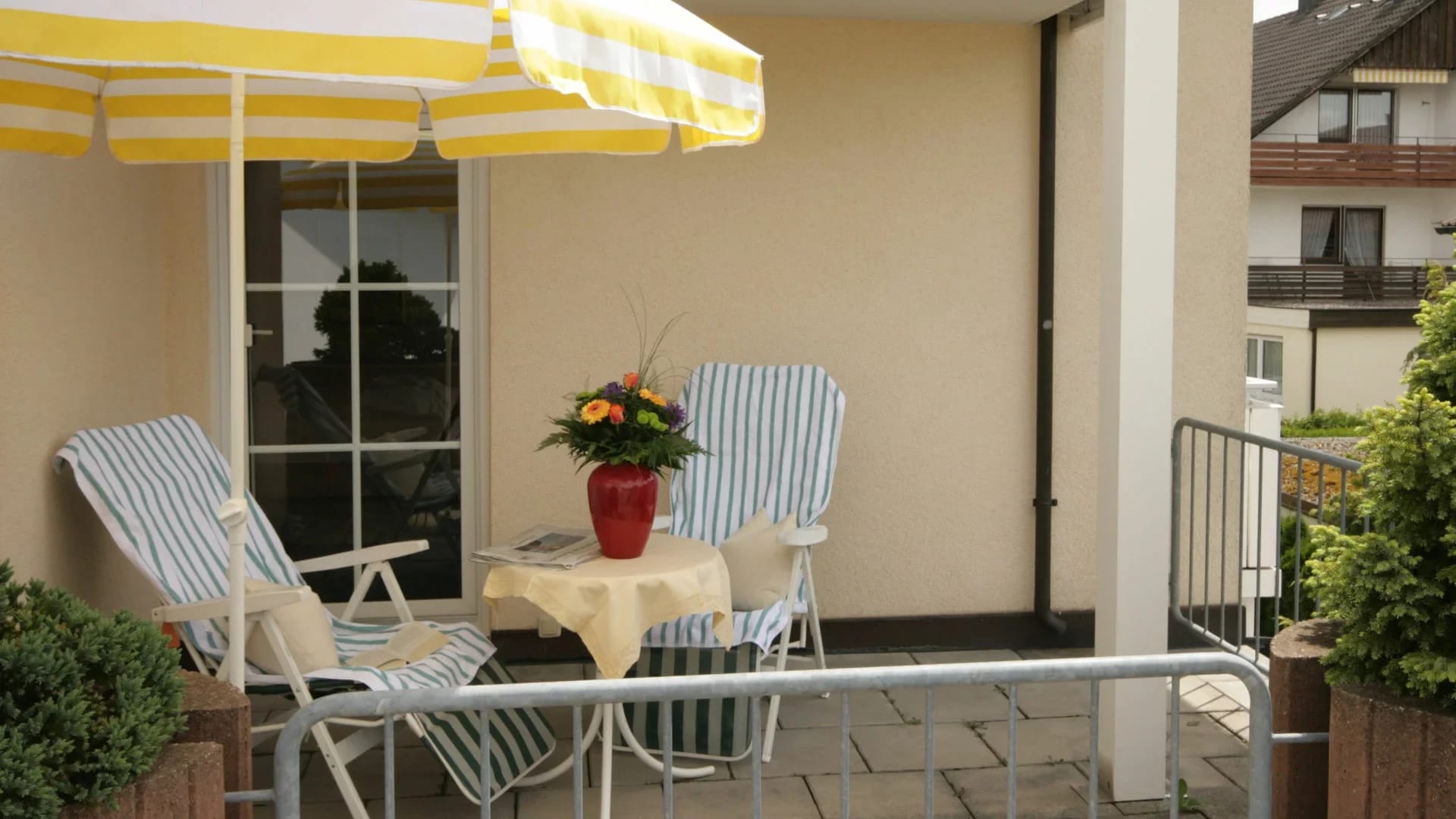 Zimmerbalkon mit gelb-weiß gestreiftem Sonnenschirm, Liegestühlen und Tisch mit Blumenstrauß