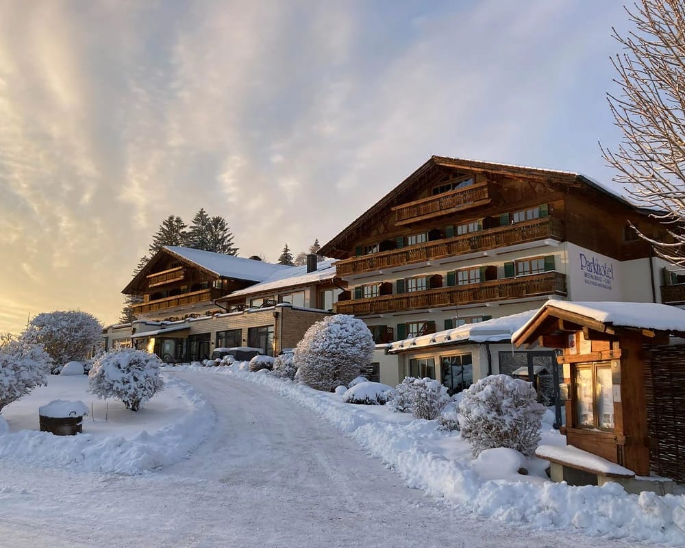 Außenansicht des Hotels mit Holzbalkonen, verschneiter Zufahrt und beleuchtetem Eingang in der Abenddämmerung