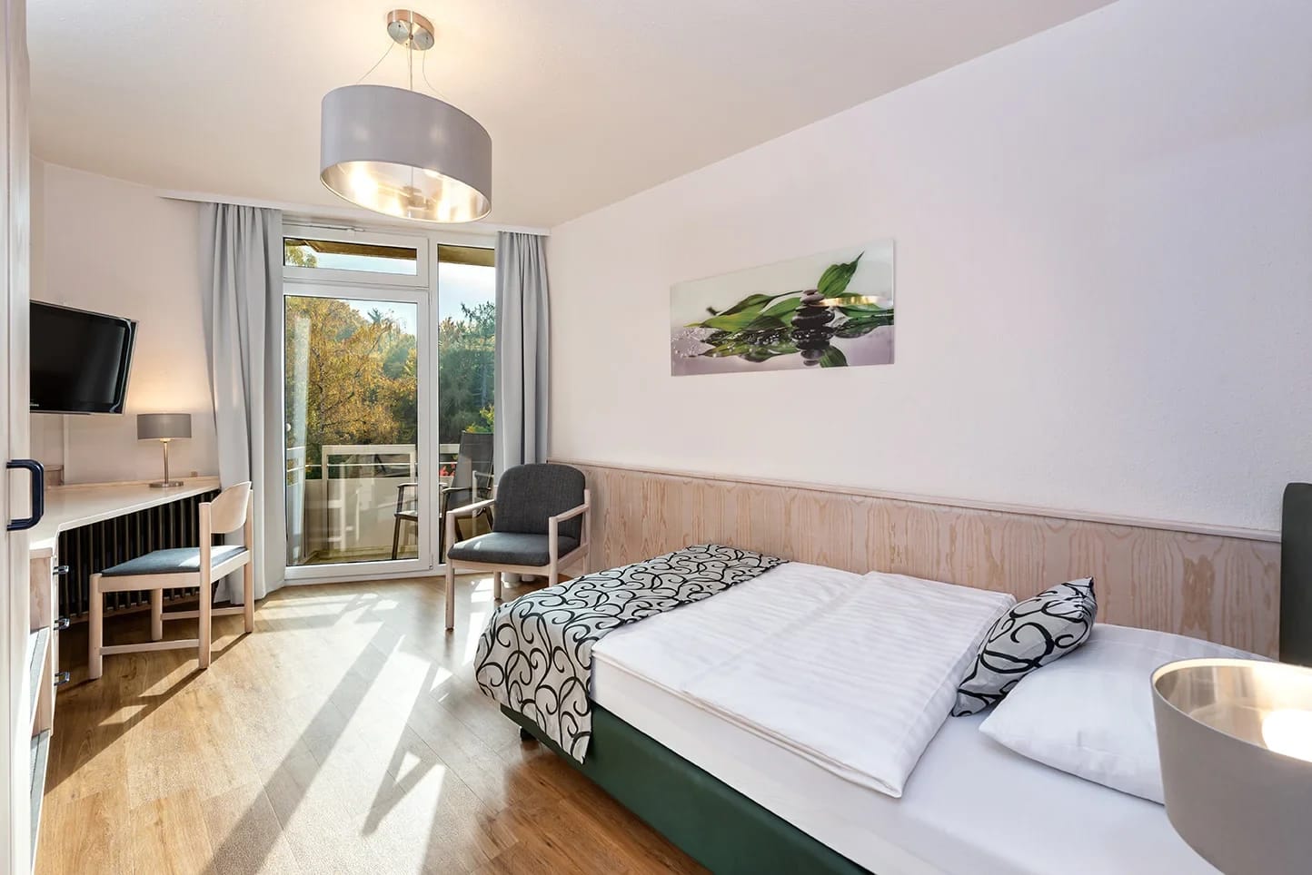 Helles Zimmer mit Bett, Schreibtisch, Fernseher und Balkontür mit Blick ins Grüne