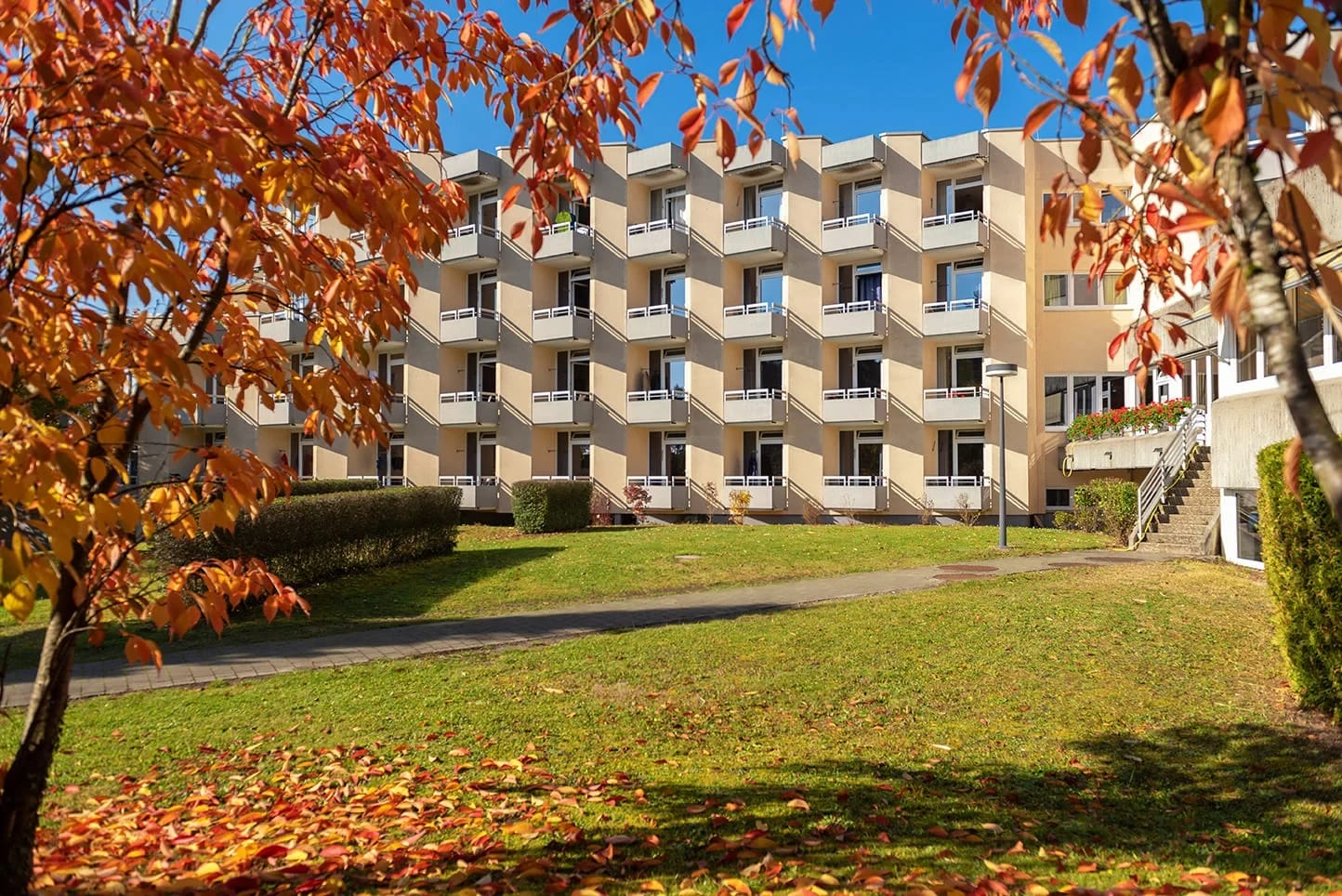 Außenansicht des Klinikgebäudes mit Balkonreihen, umrahmt von herbstlich gefärbten Bäumen und Rasenfläche