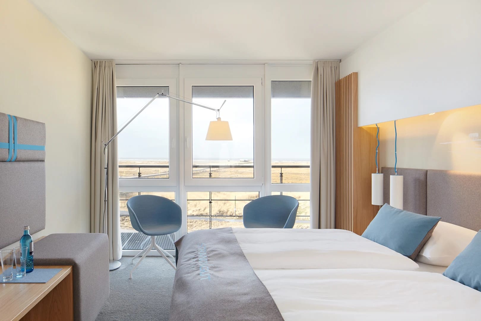 Zimmer mit zwei Betten, blauen Sesseln und bodentiefen Fenstern mit Blick über die Dünen