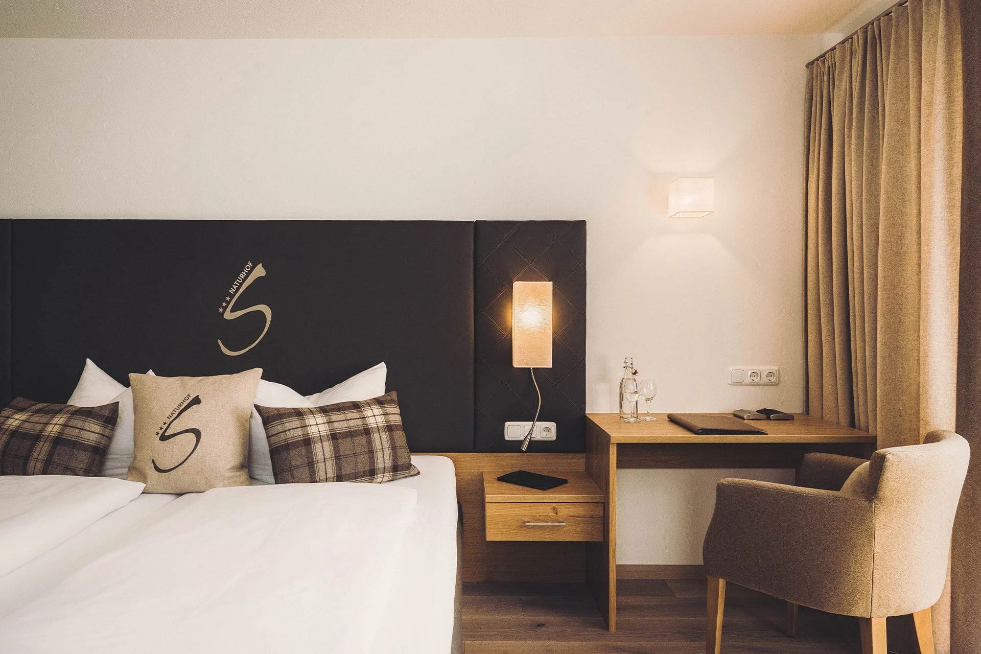 Zimmer mit Doppelbett, dunklem Polsterkopfteil, Schreibtisch und Sessel