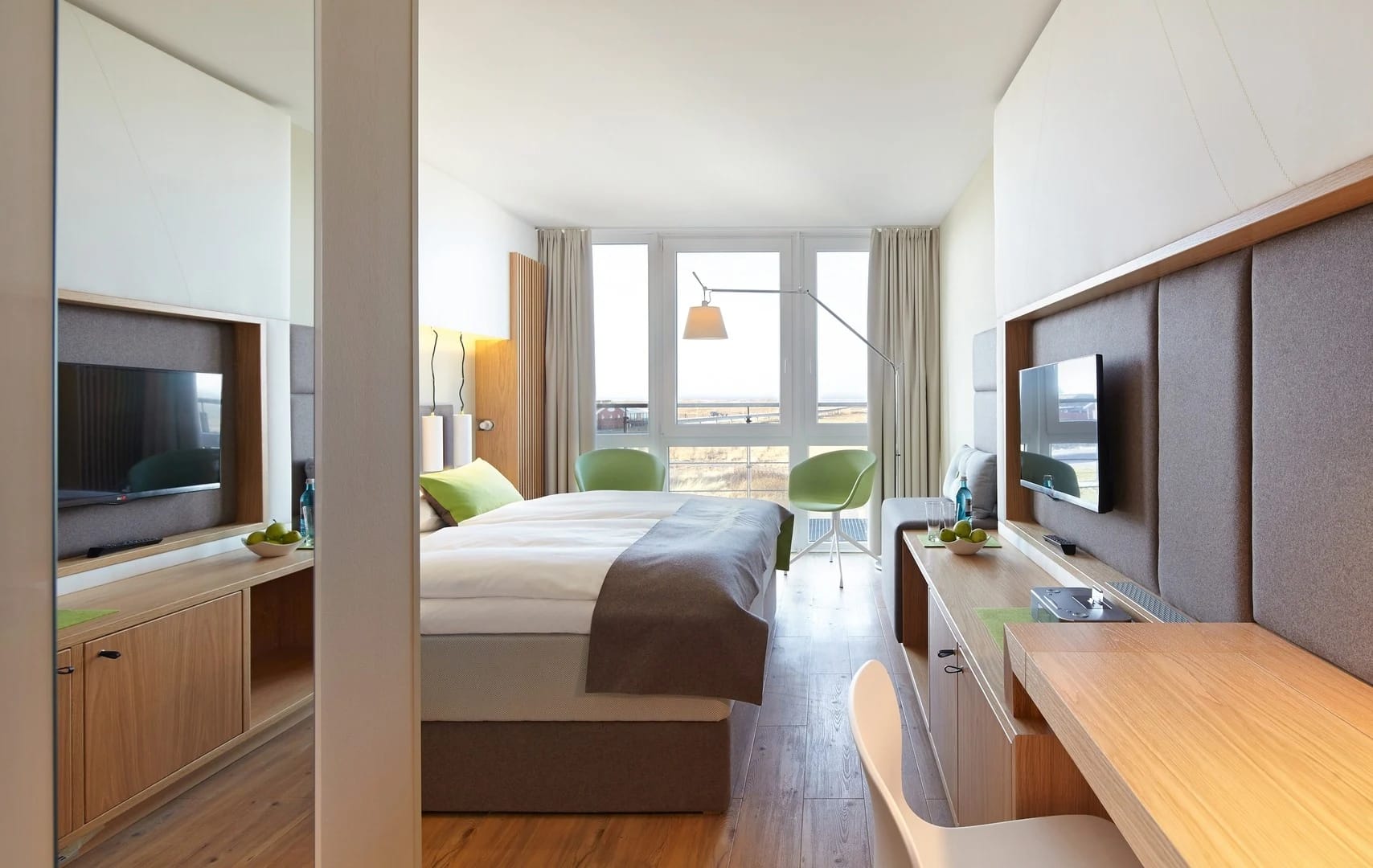 Helles Zimmer mit Doppelbett, Schreibtisch, Fernseher und grünen Sesseln vor bodentiefem Fenster