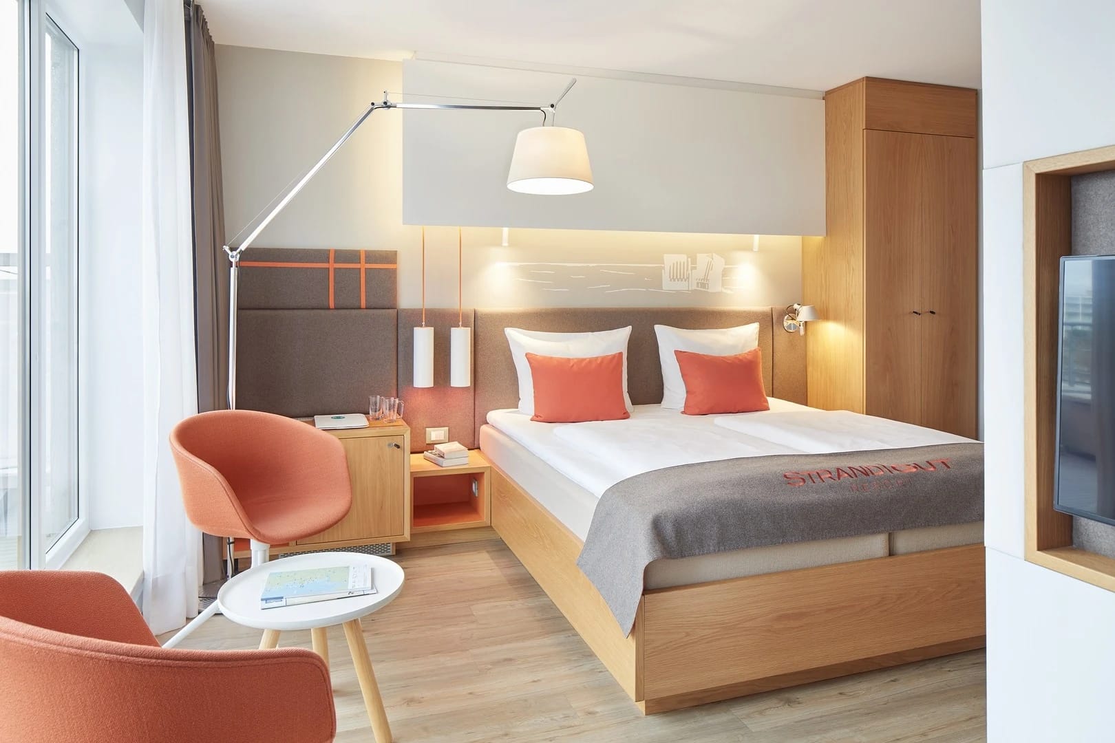 Zimmer mit Doppelbett, orangefarbenen Sesseln und Schreibtisch am Fenster