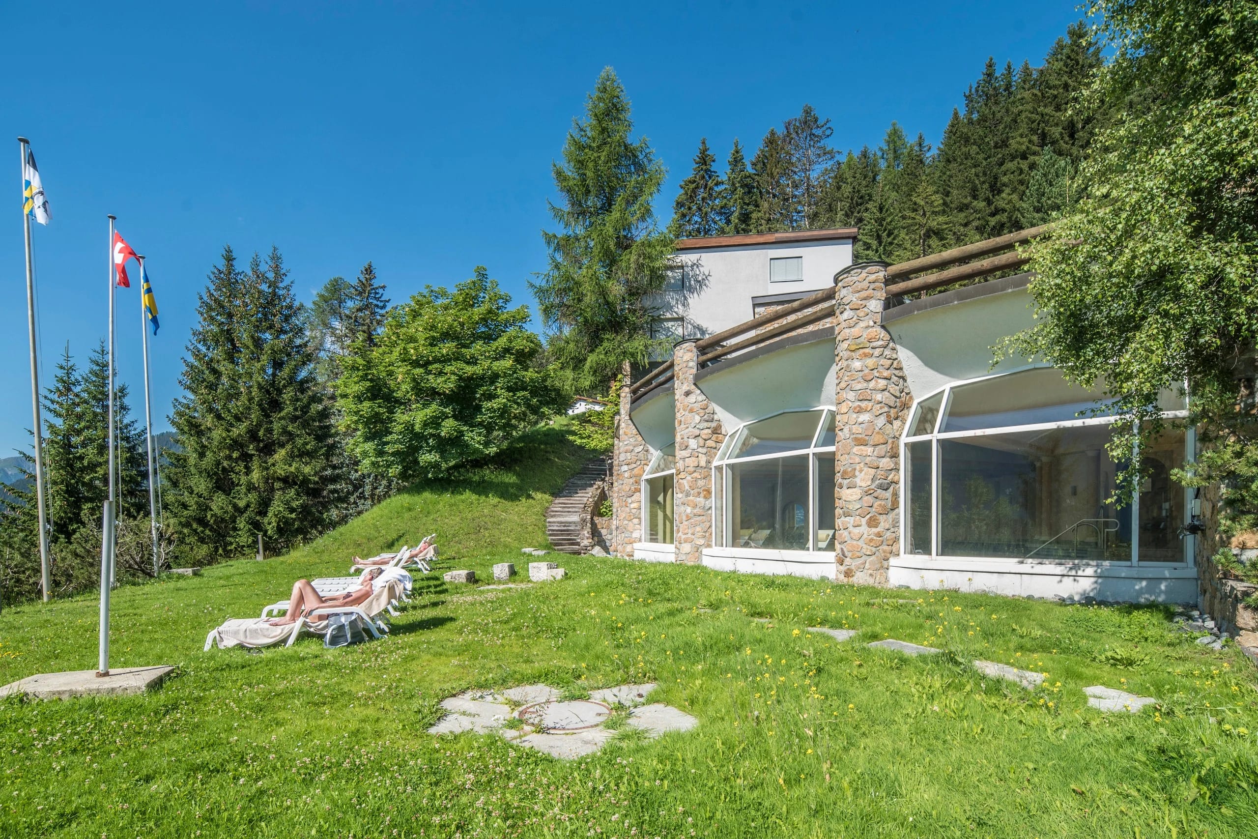 Wellnessgebäude mit Natursteinsäulen und Glasfront, davor Liegewiese mit Sonnenliegen und Fahnenmasten