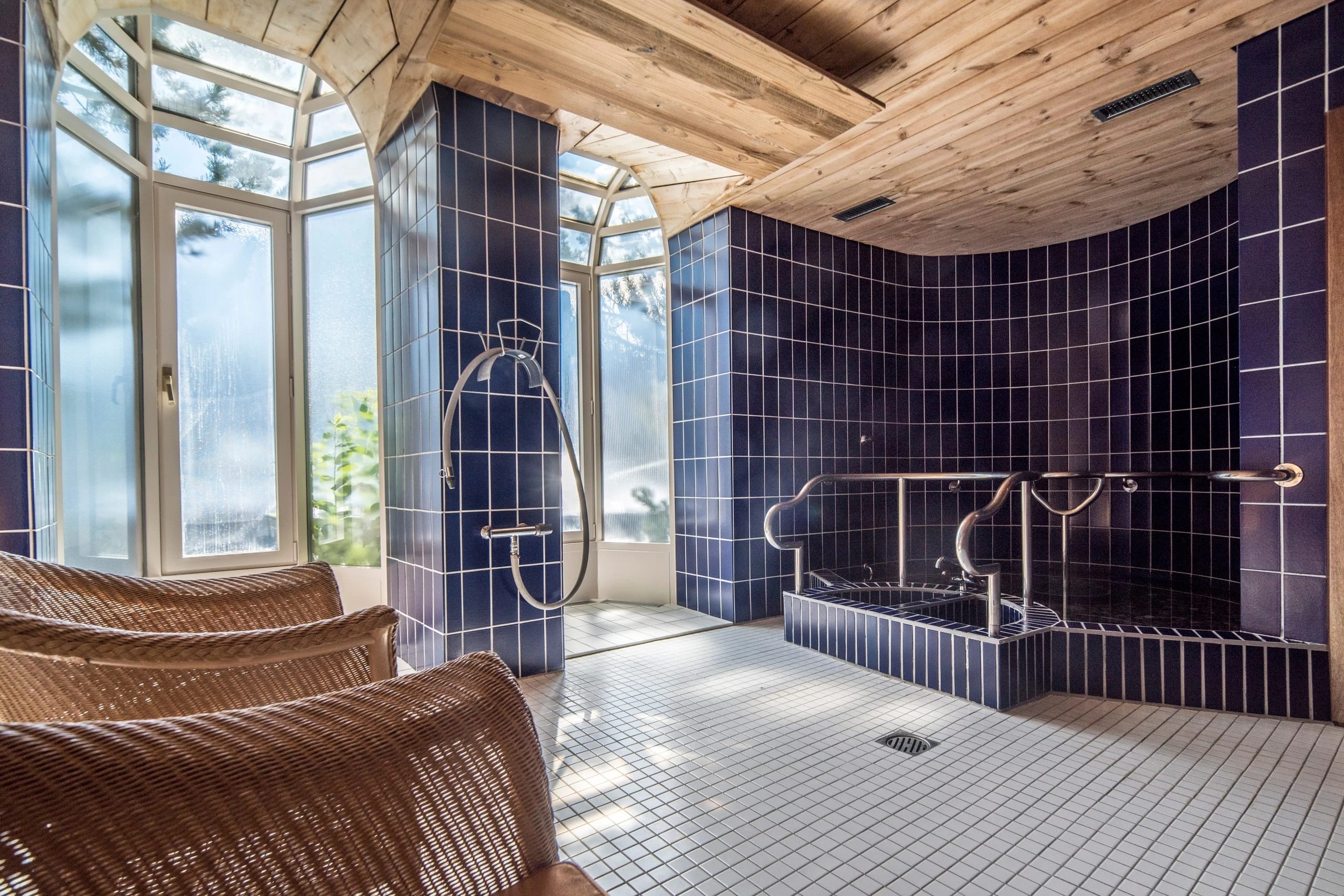 Wellnessraum mit blau gefliestem Whirlpool, Erlebnisdusche und Korbliegen unter Holzdecke