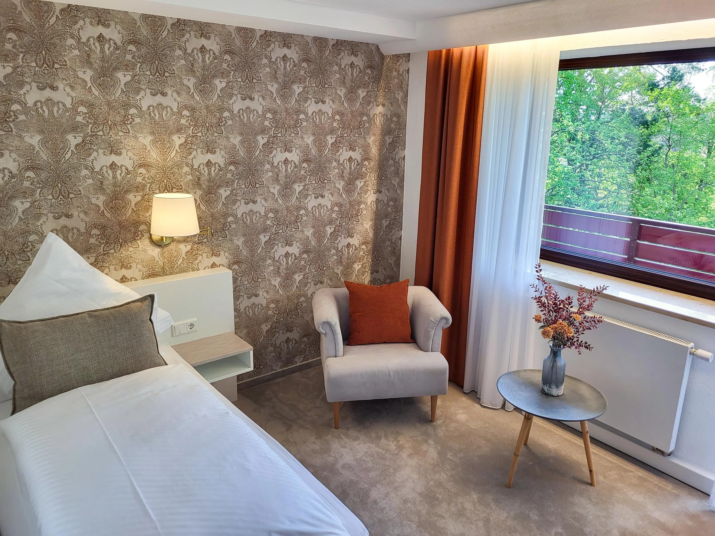 Zimmer mit gemusterter Tapete, hellem Sessel und Beistelltisch am Fenster