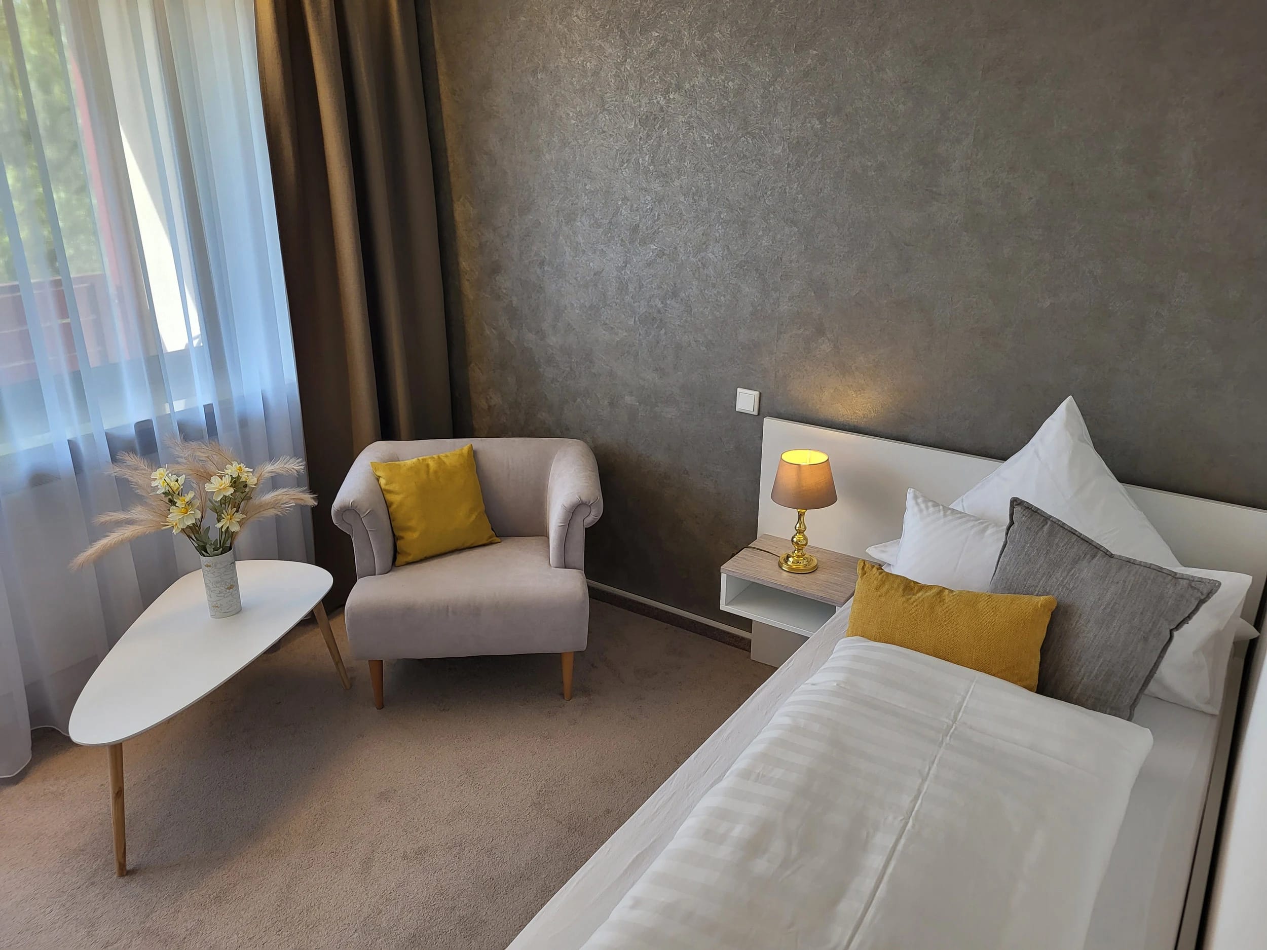 Zimmer mit Bett, gelben Kissen, hellem Sessel und kleinem Beistelltisch mit Blumen