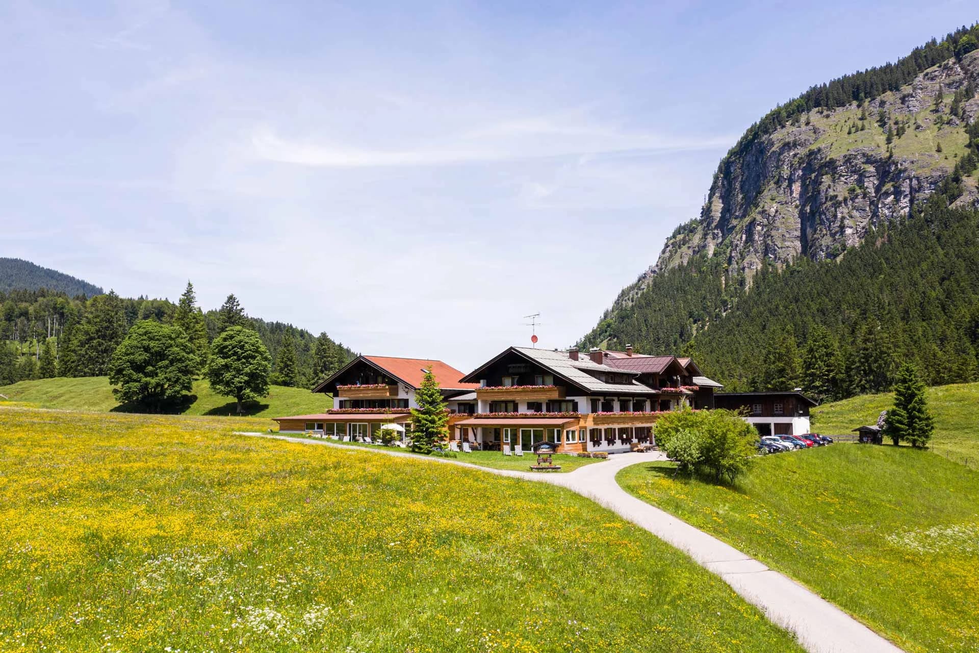 Hotelgebäude in einer blühenden Bergwiese, dahinter Wald und eine steile Felswand