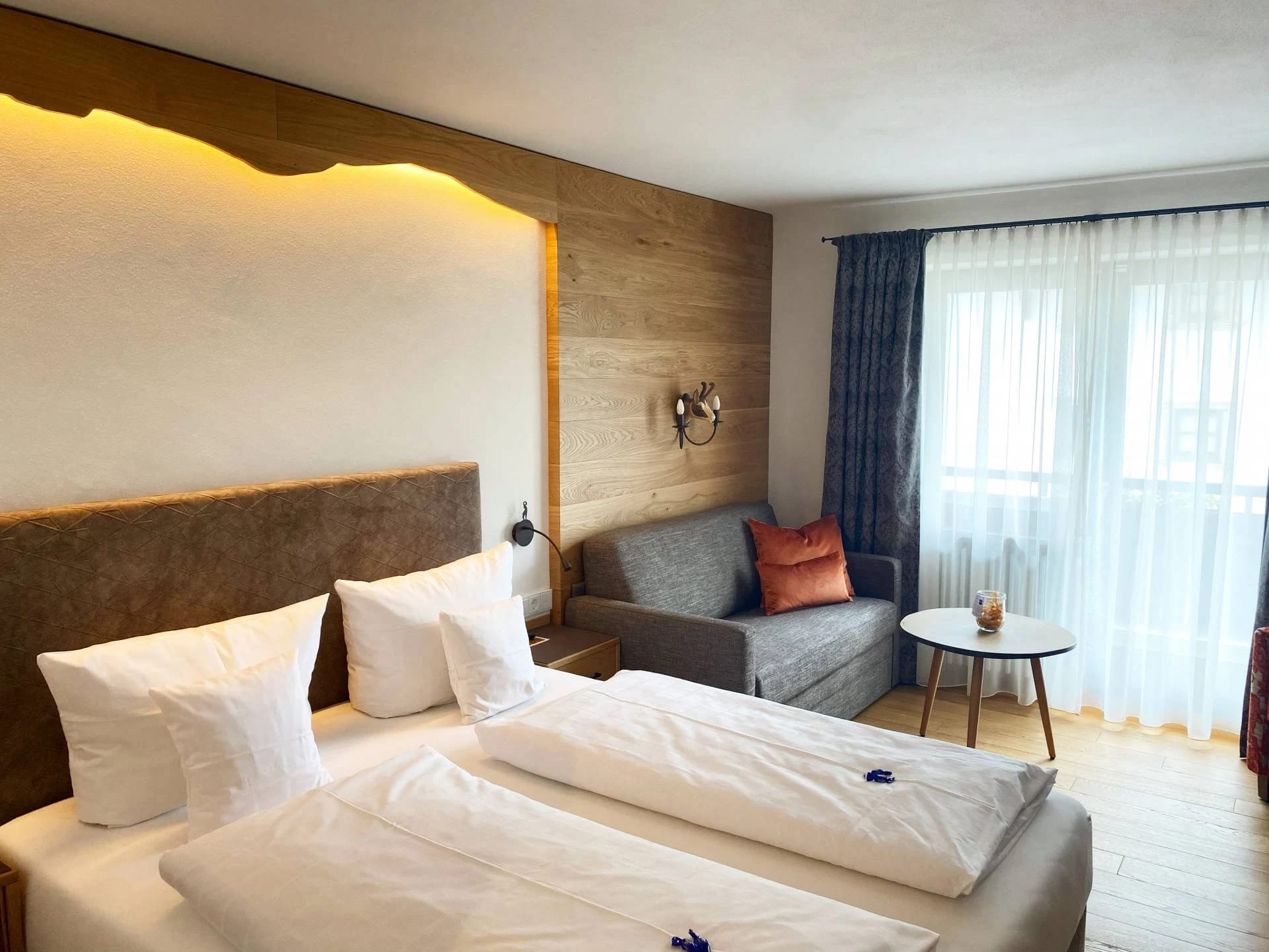 Helles Zimmer mit Doppelbett vor Holzwand, Sofa, rundem Beistelltisch und bodenlangem Vorhang