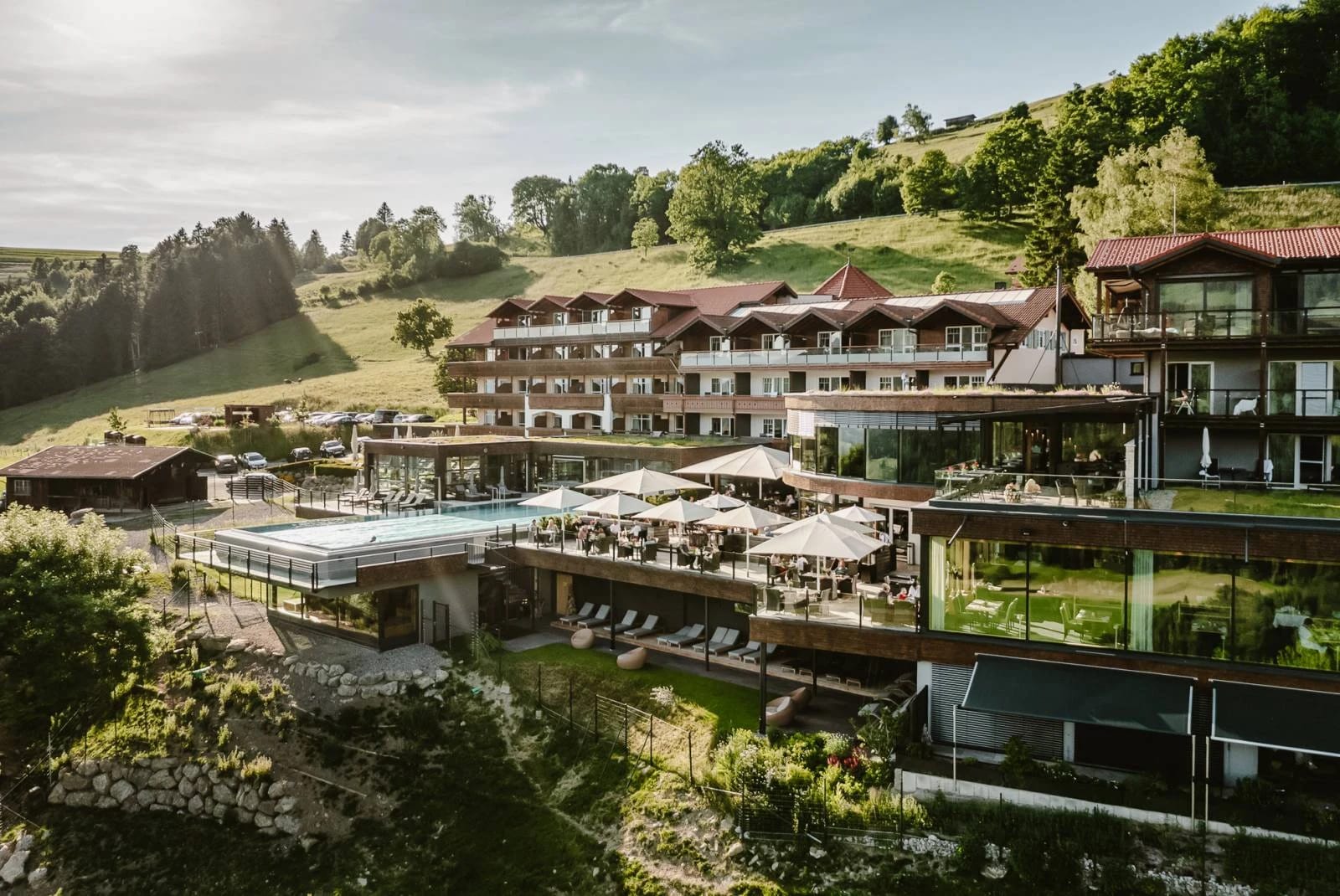 Luftaufnahme des Resort Bergkristall mit Außenpool und Sonnenschirmen vor grünen Hügeln