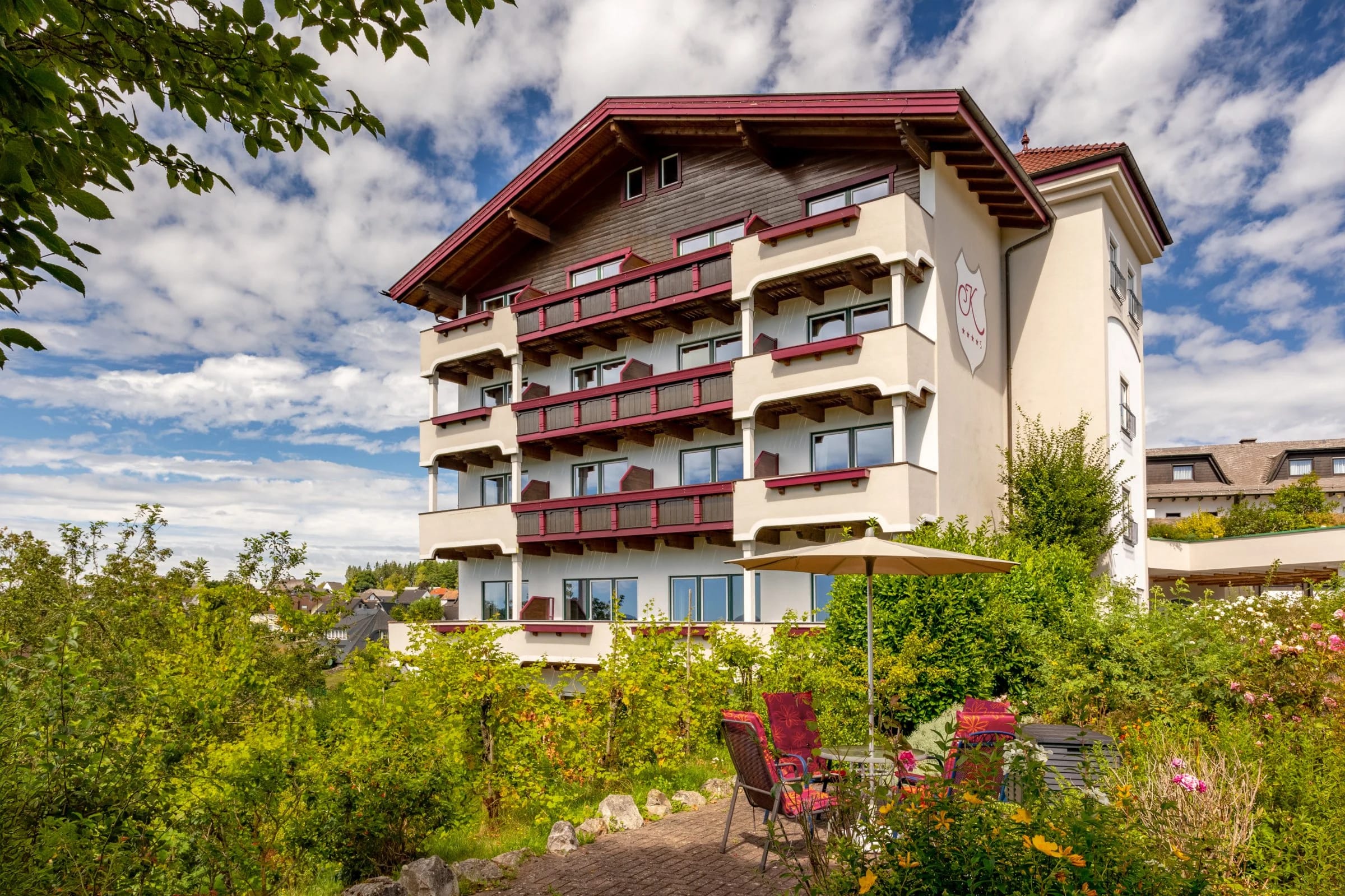 Hotelgebäude mit Holzbalkonen, davor Gartenterrasse mit Stühlen und Sonnenschirm