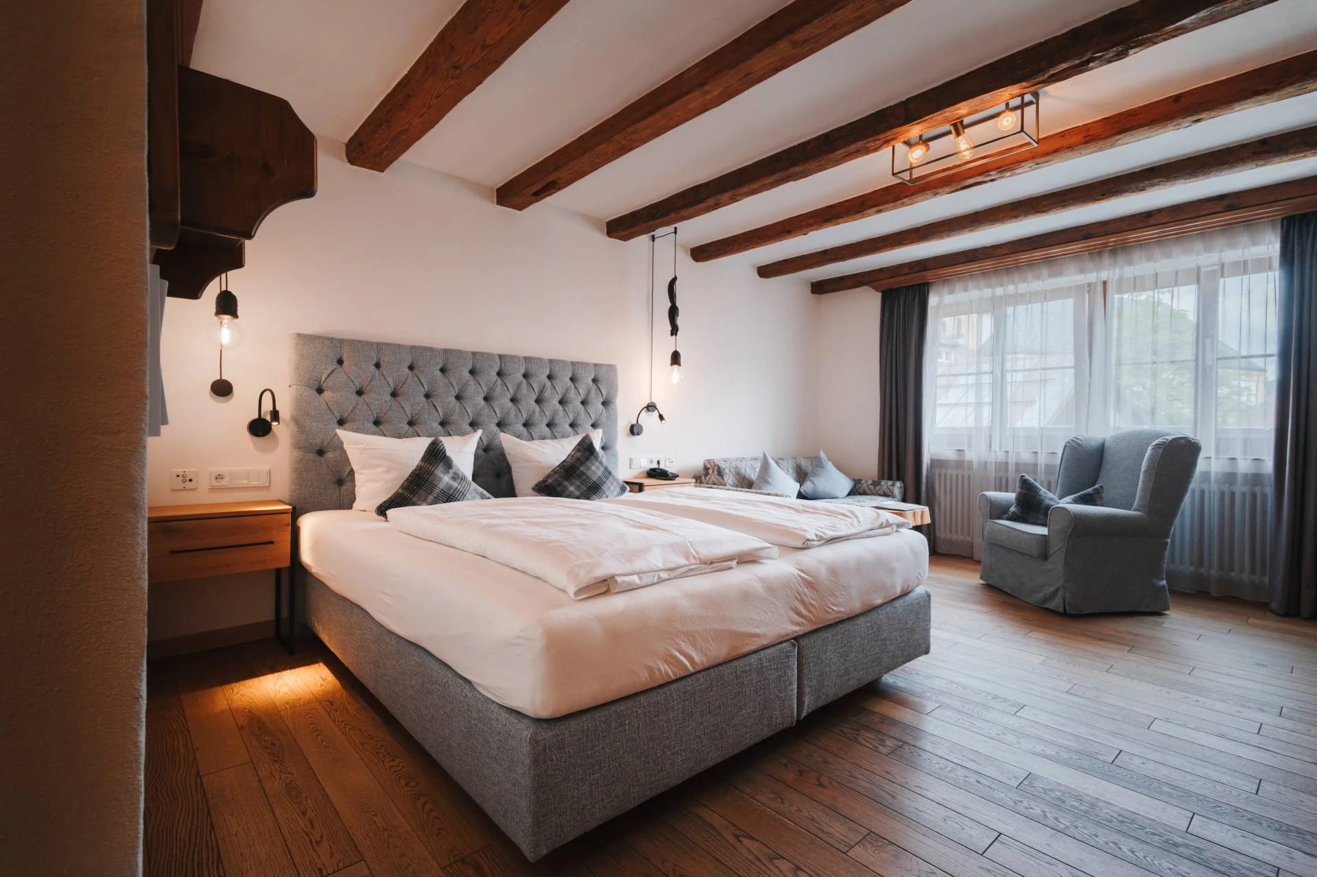 Zimmer mit Holzbalkendecke, Doppelbett mit gepolstertem Kopfteil, Sessel und breiter Fensterfront