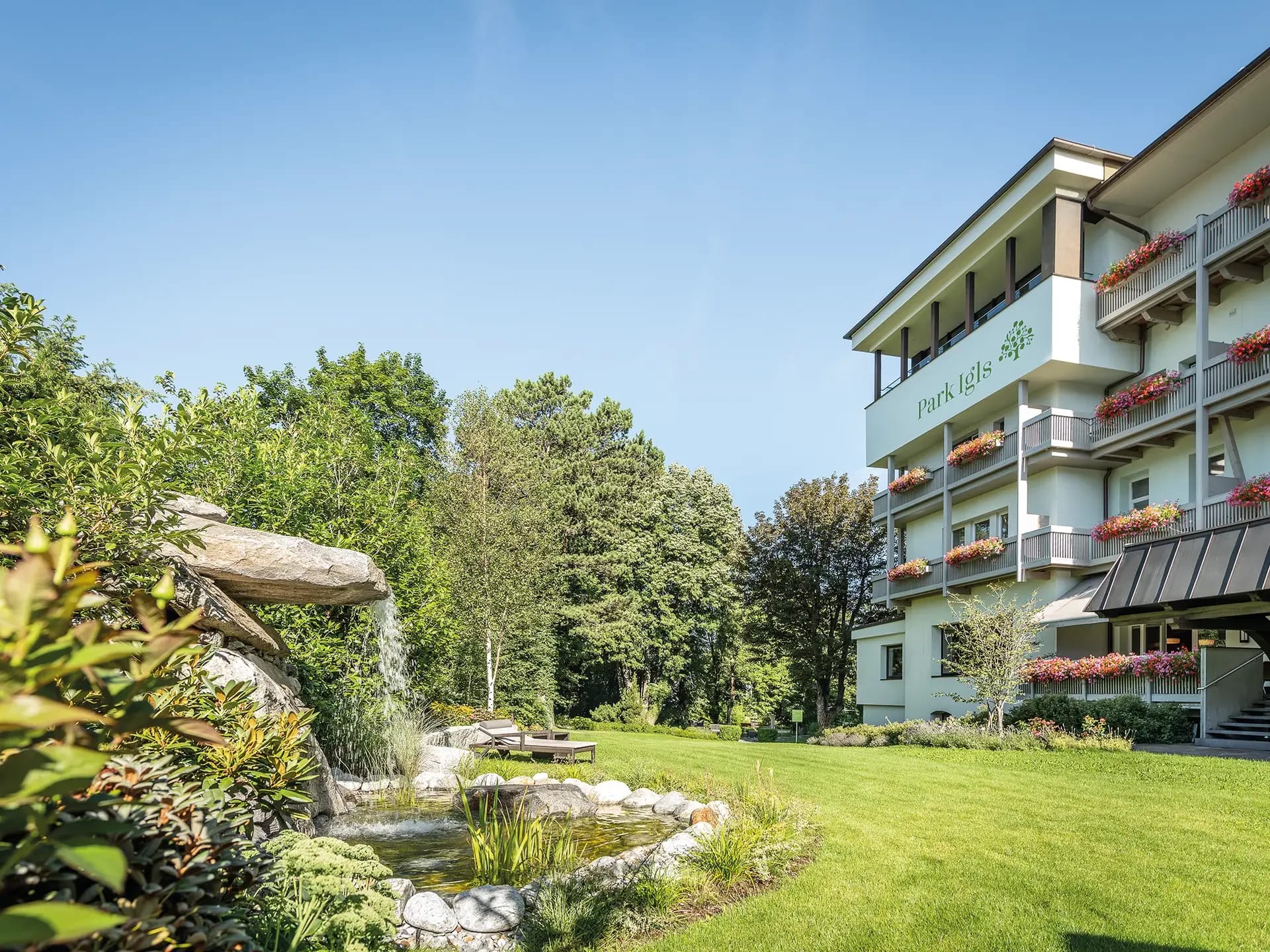 Gartenanlage mit kleinem Teich und Wasserlauf vor dem Hotelgebäude mit blumengeschmückten Balkonen