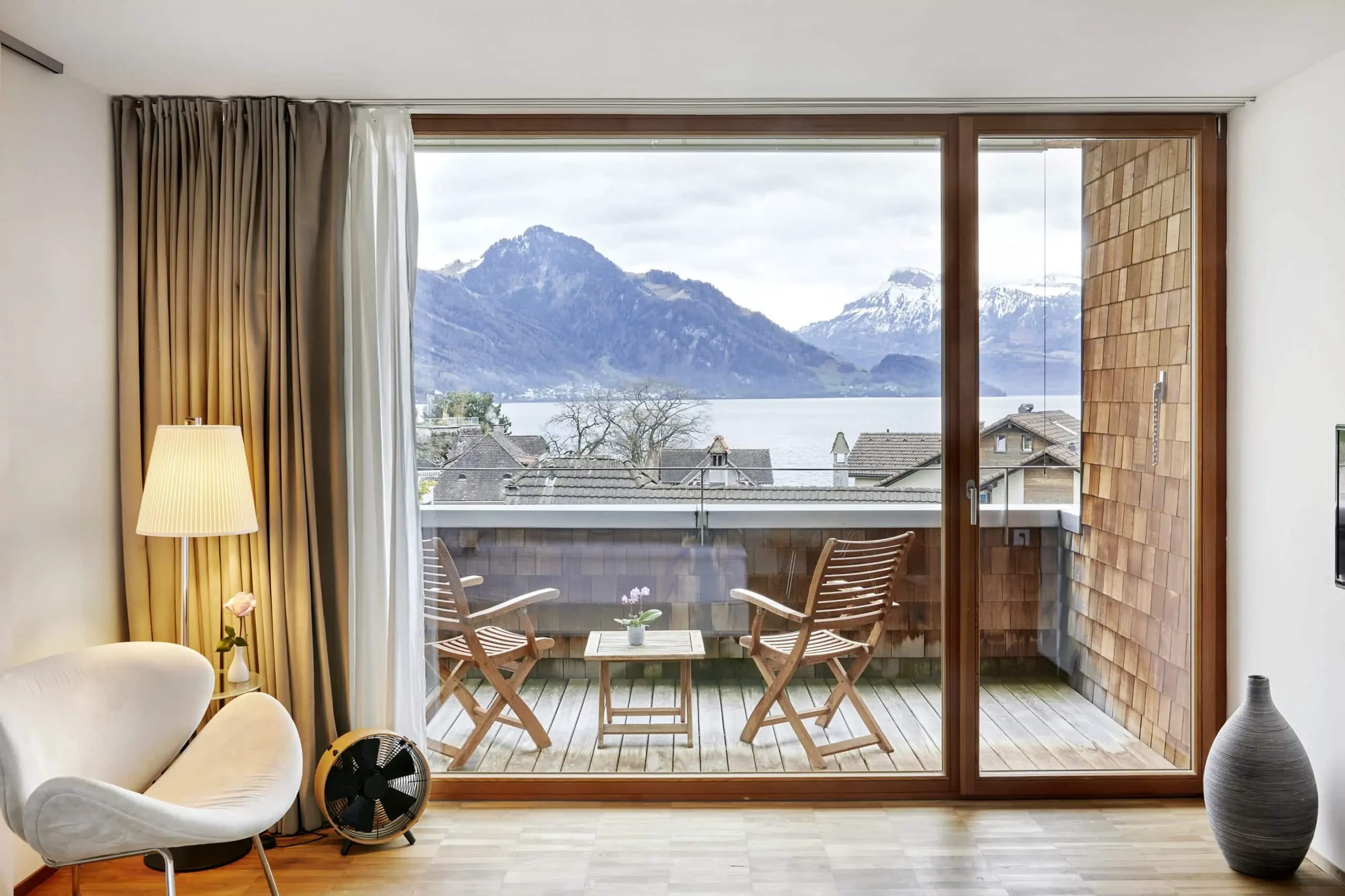 Zimmer mit raumhoher Fensterfront und Balkon mit Holzstühlen, Blick auf See und verschneite Berge