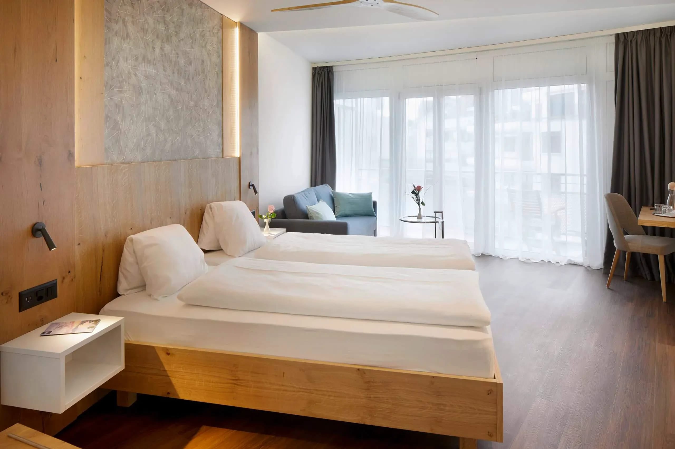 Zimmer mit Holzwand hinter dem Doppelbett, Sofa und bodentiefer Fensterfront zum Balkon