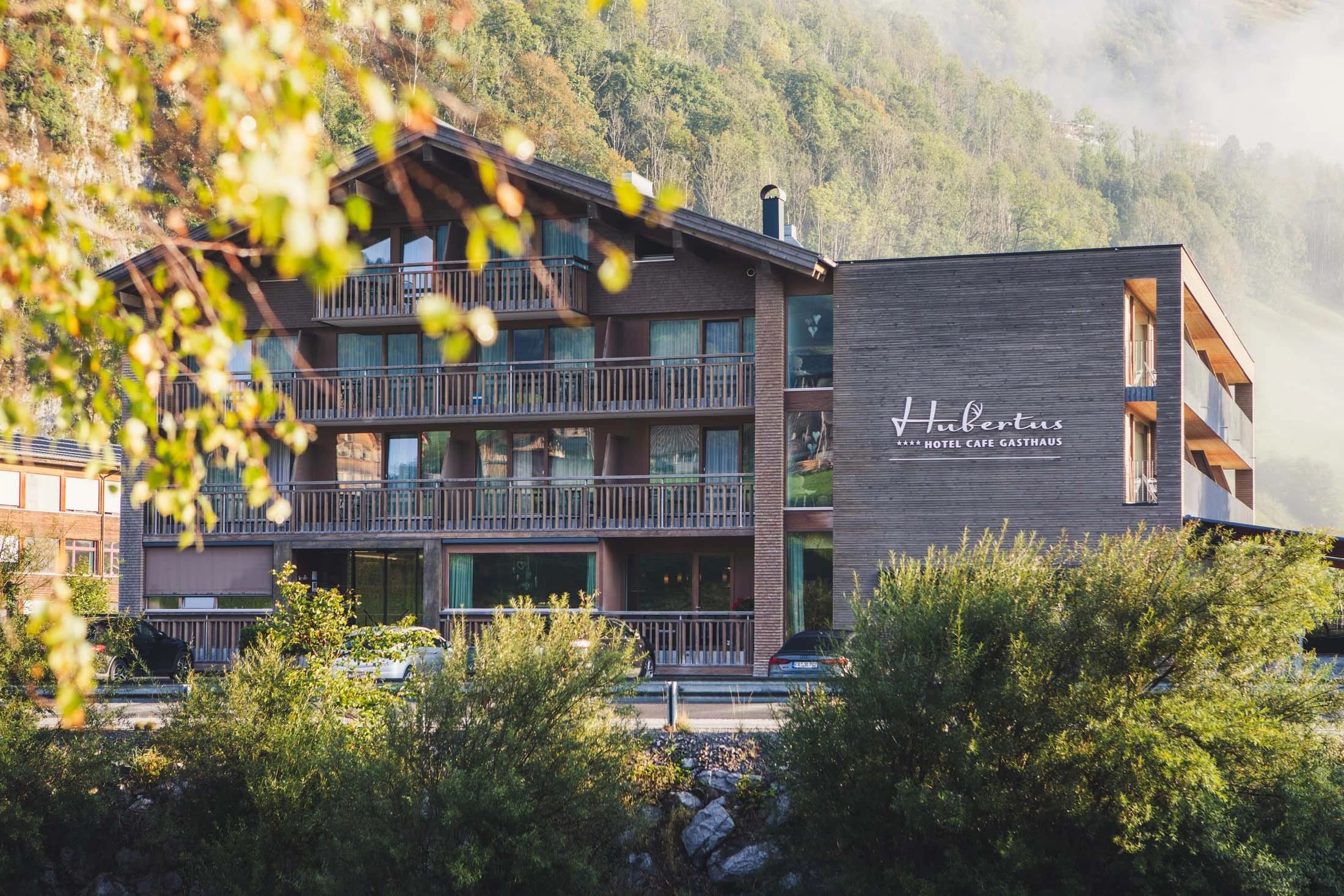Außenansicht des Hotels mit Holzfassade und Balkonen vor bewaldetem Berghang, im Vordergrund herbstliches Laub
