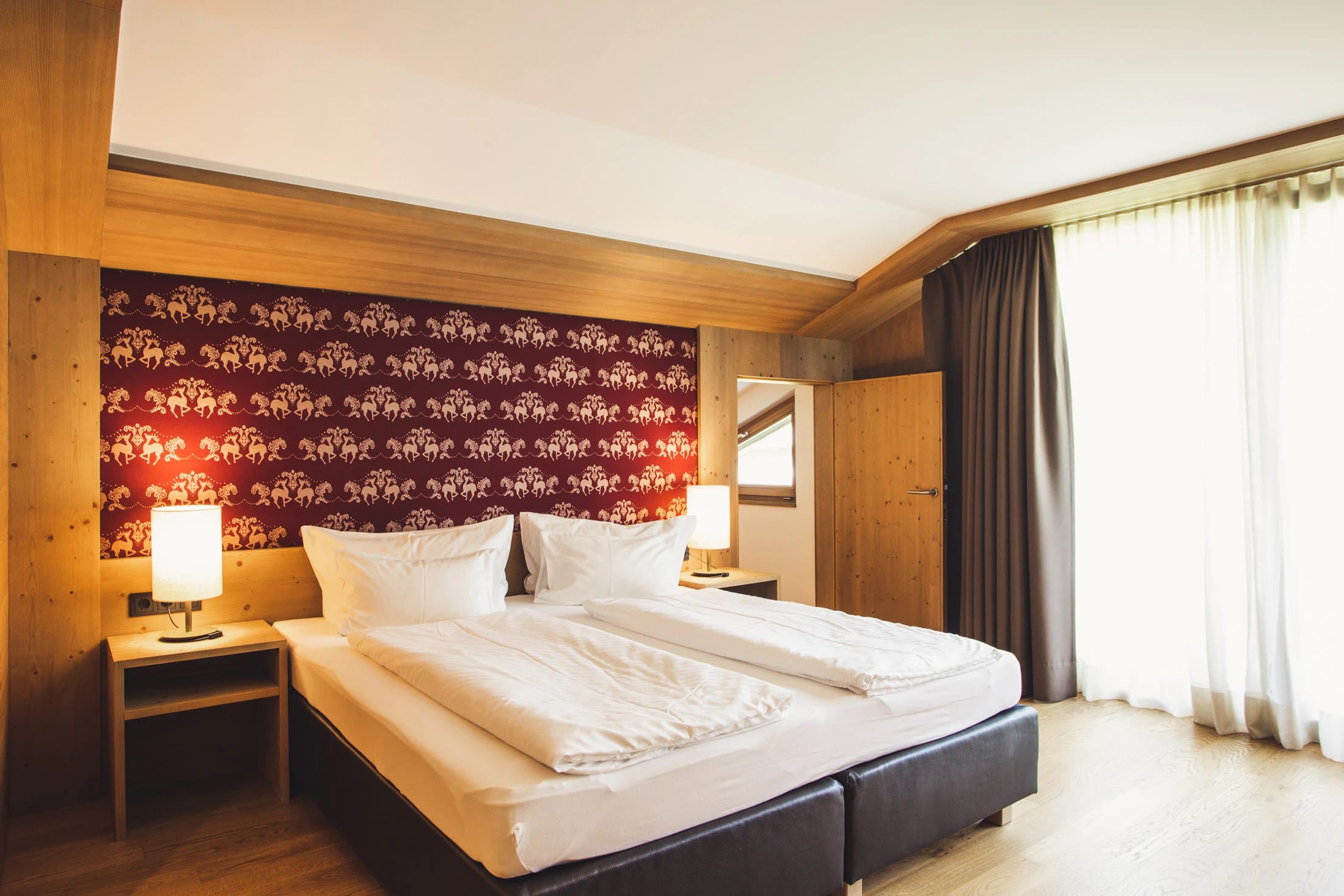Doppelzimmer mit weißer Bettwäsche vor rot gemusterter Wand, Holzverkleidung und Fenster