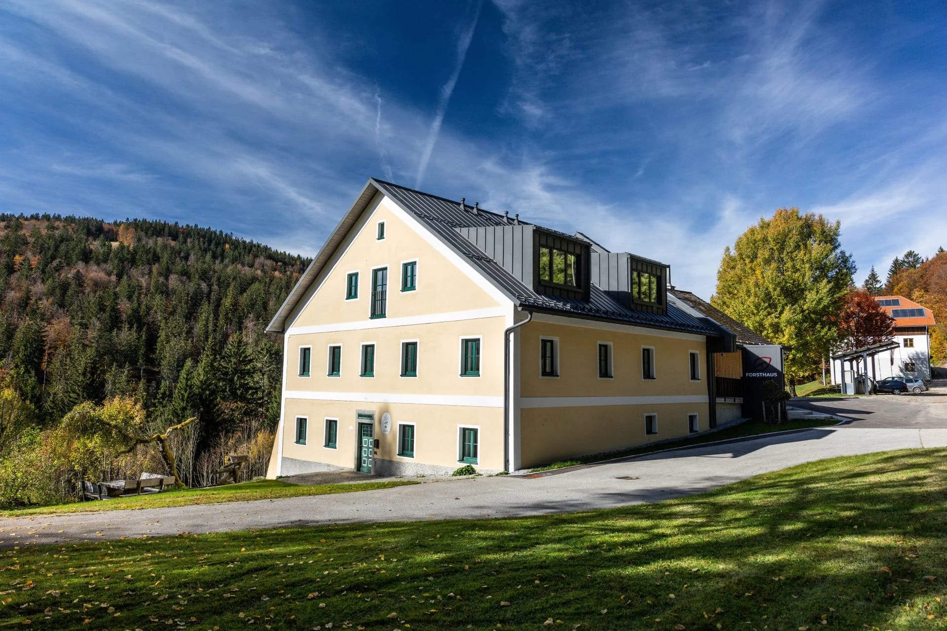 Freistehendes Gebäude mit hellgelber Fassade und dunklem Dach auf einer Wiese vor bewaldetem Hügel