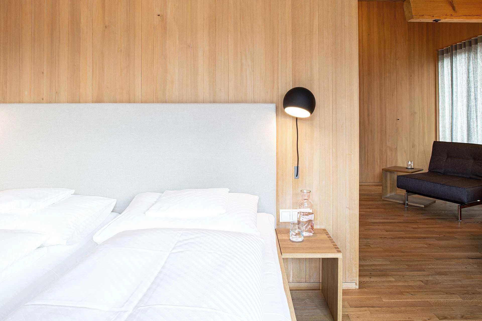 Zimmer mit Doppelbett vor heller Holzwand, schwarzer Wandleuchte und Sofa am Fenster