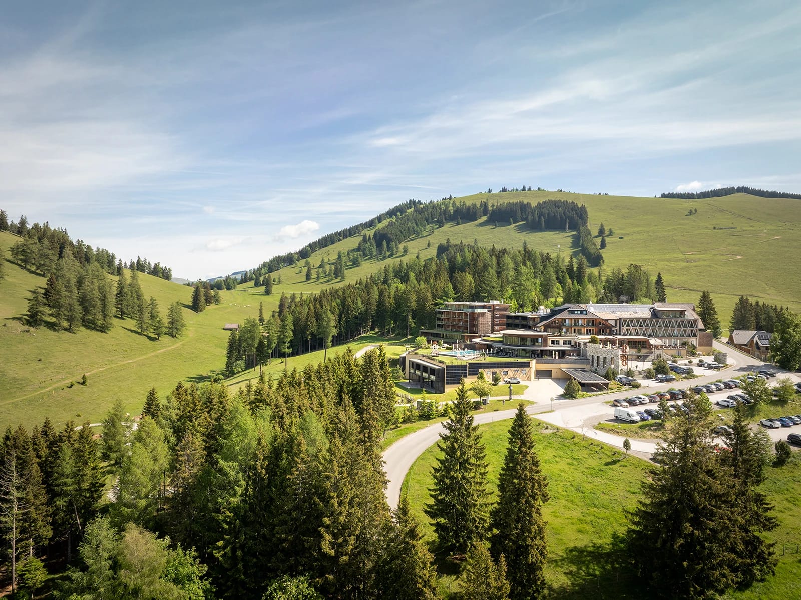 Blick über Nadelwald und grüne Almwiesen auf das Hotel in der Landschaft