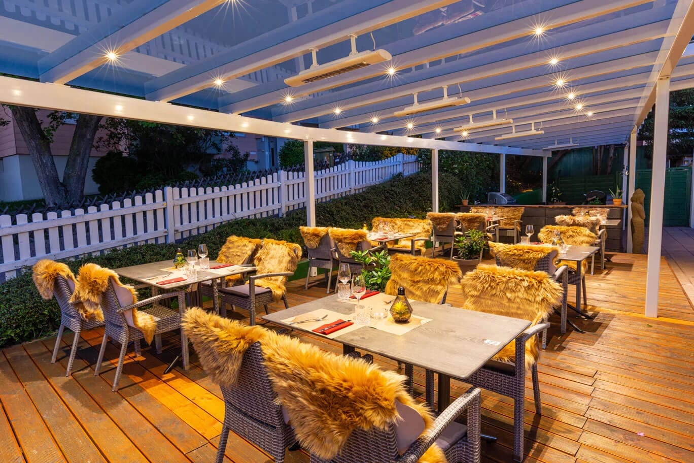 Überdachte Holzterrasse am Abend mit gedeckten Tischen, Korbstühlen mit Felldecken und weißem Lattenzaun