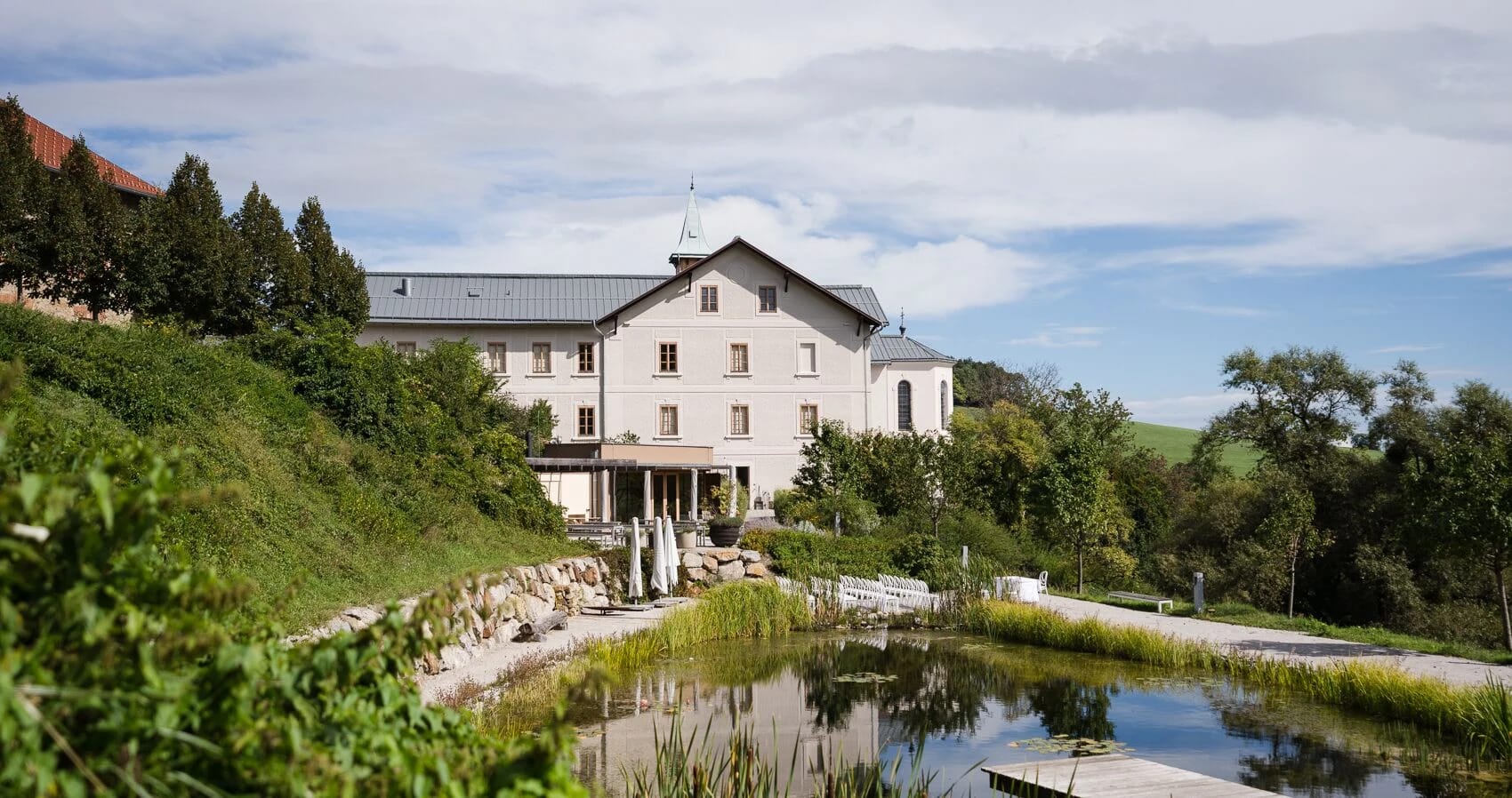 Weißes Haupthaus mit Turm auf einem grünen Hang, davor ein Naturteich mit Uferbewuchs